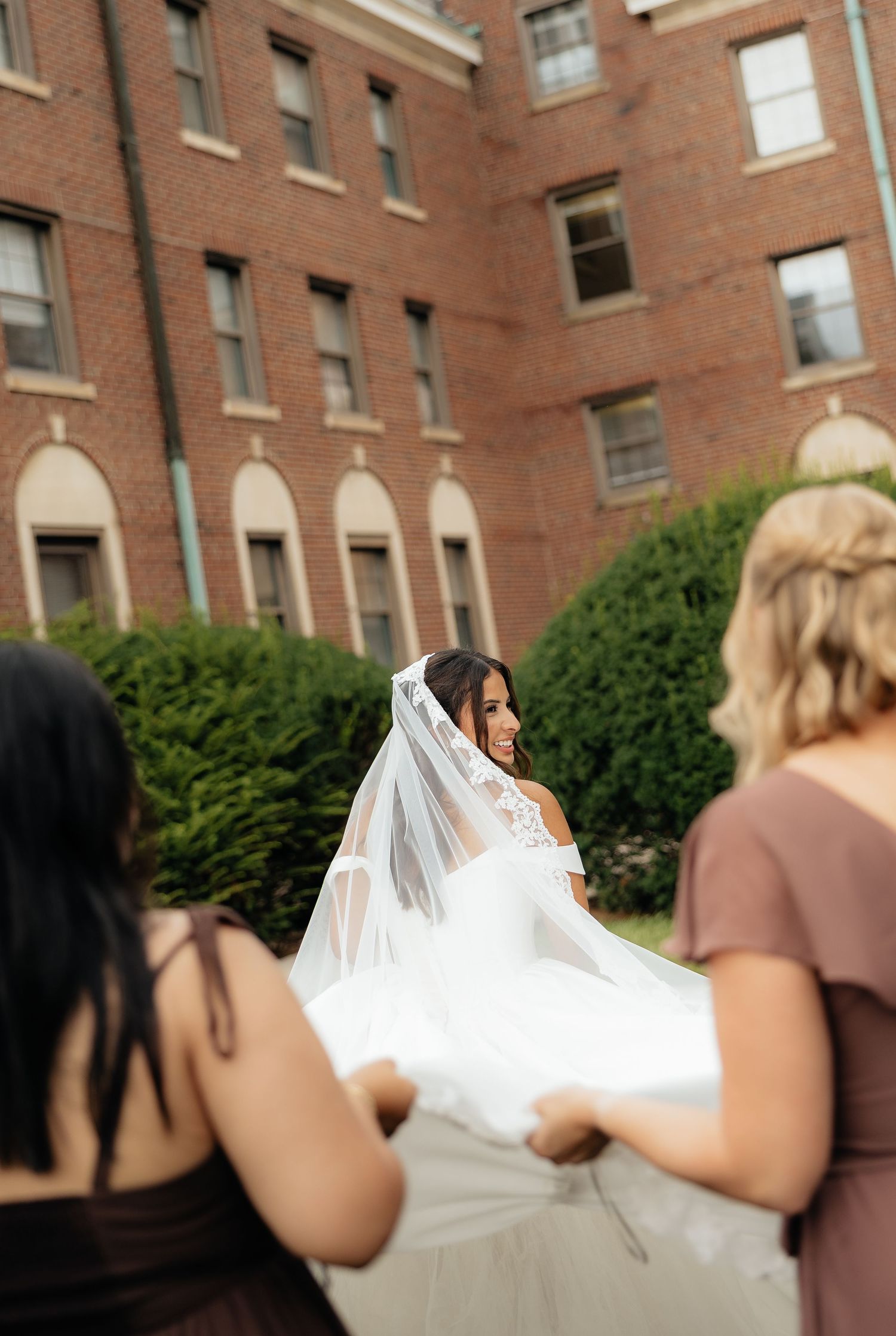 Bride in white gown and veil smiles as bridesmaids help carry her dress outside a brick building.