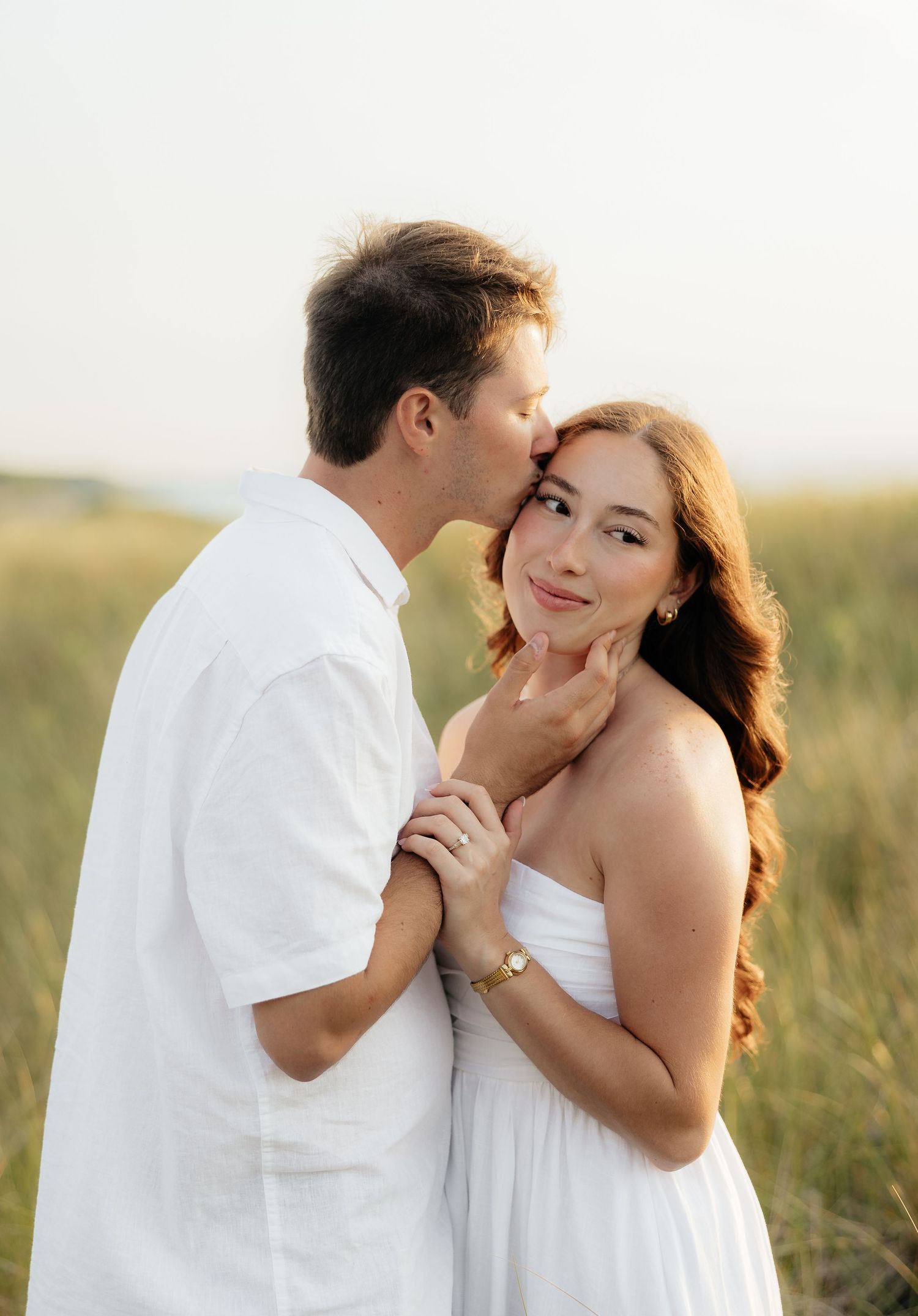 A couple in white outfits share a romantic moment in a golden grass field, man kissing woman's cheek.