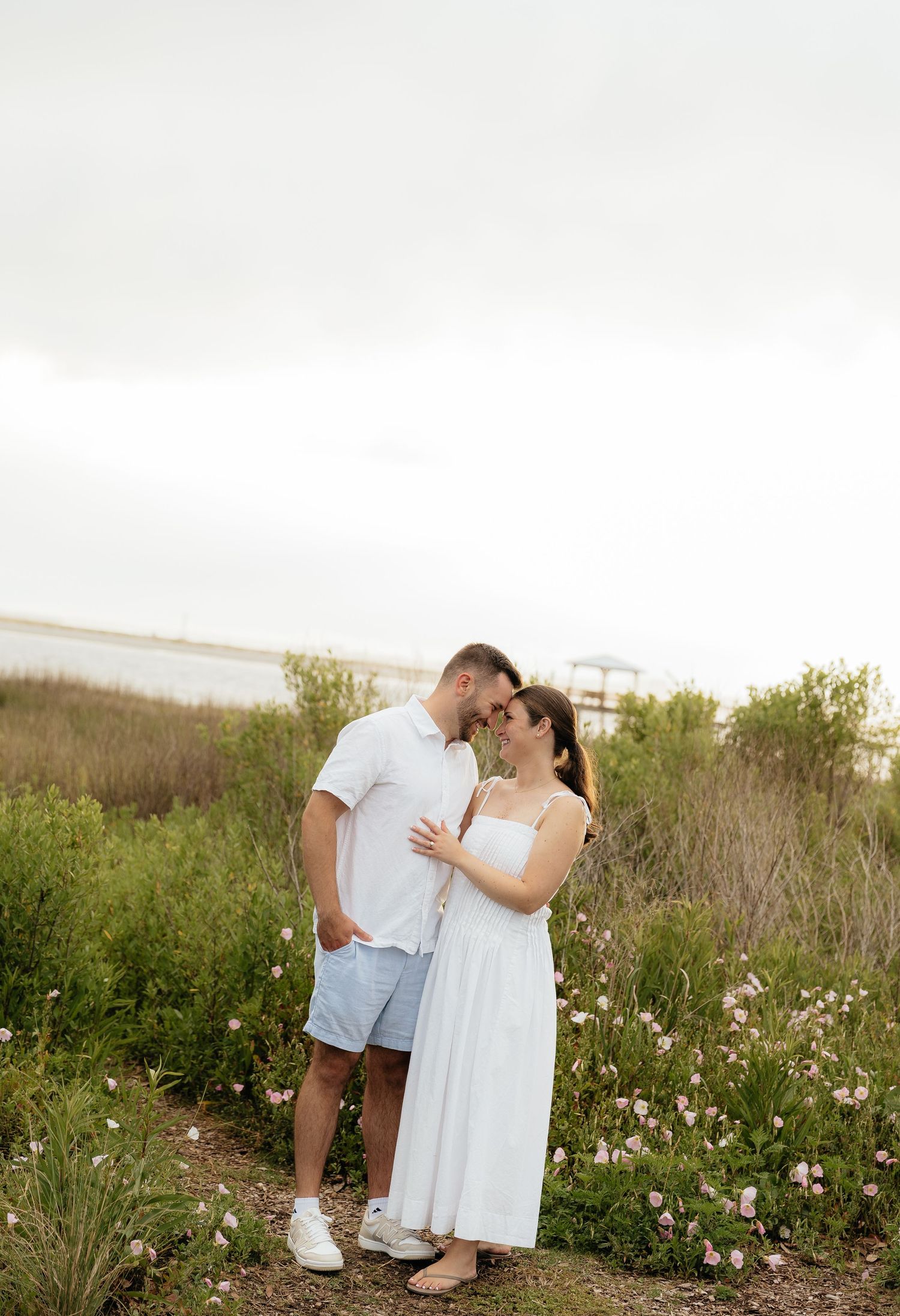 Couple in white outfits embracing among wildflowers near a coastal waterway on a cloudy day.