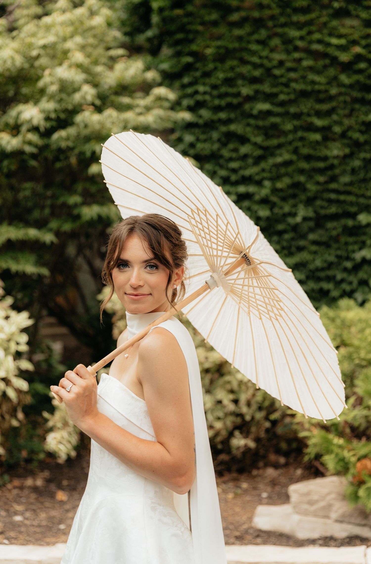 Bride in white dress poses with parasol against ivy covered wall.
