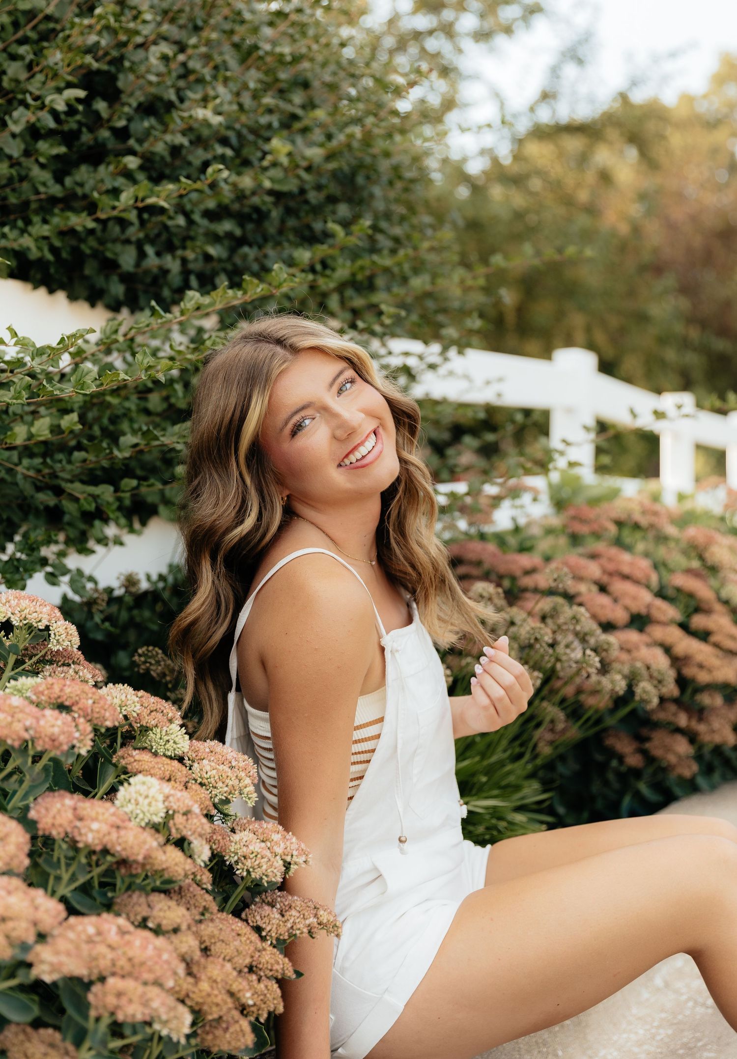 A figure in a white sleeveless top poses gracefully outdoors against a backdrop of pink hydrangea flowers and greenery.