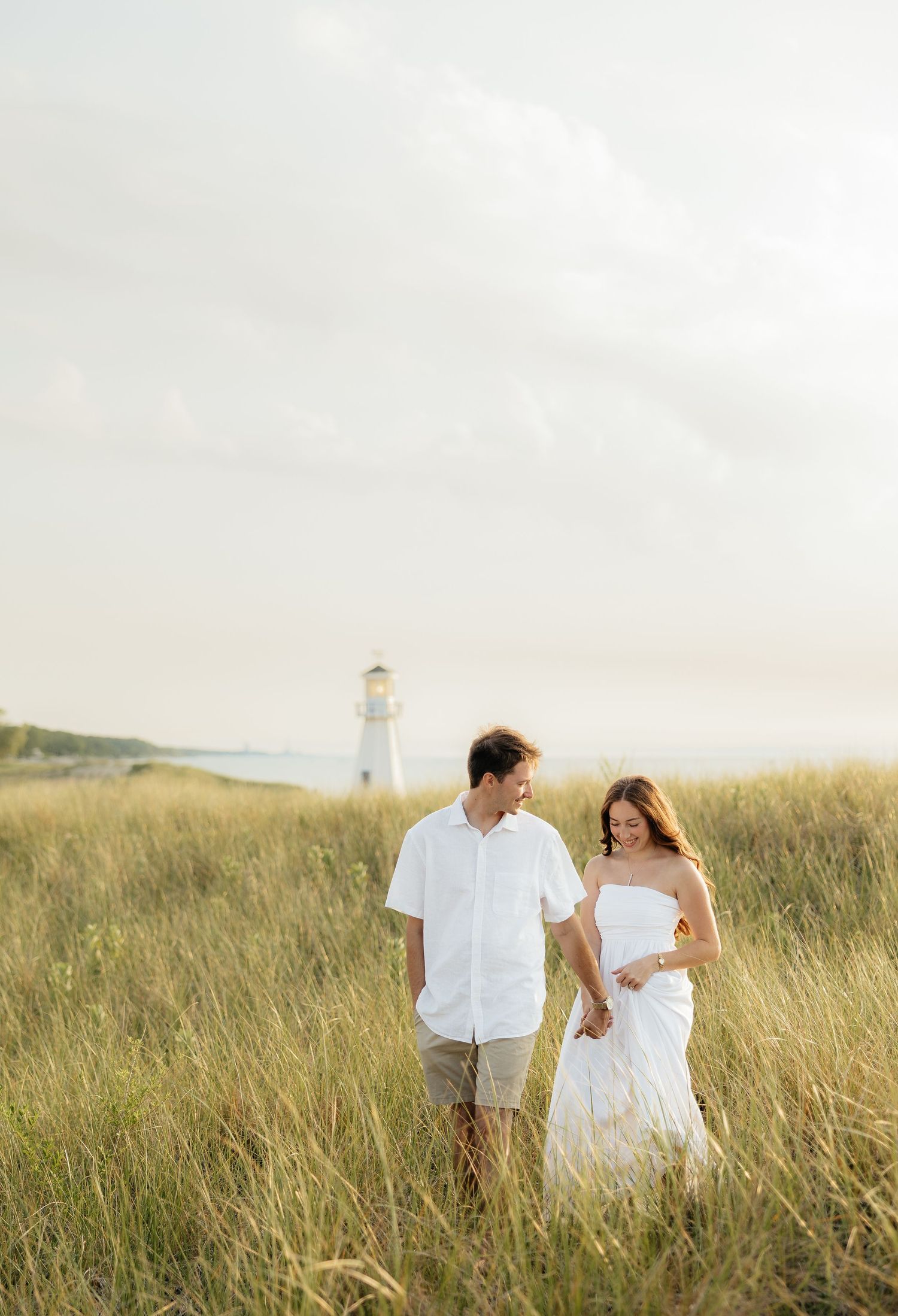 Couple in white outfits walking hand in hand through tall grass with a lighthouse in the background.