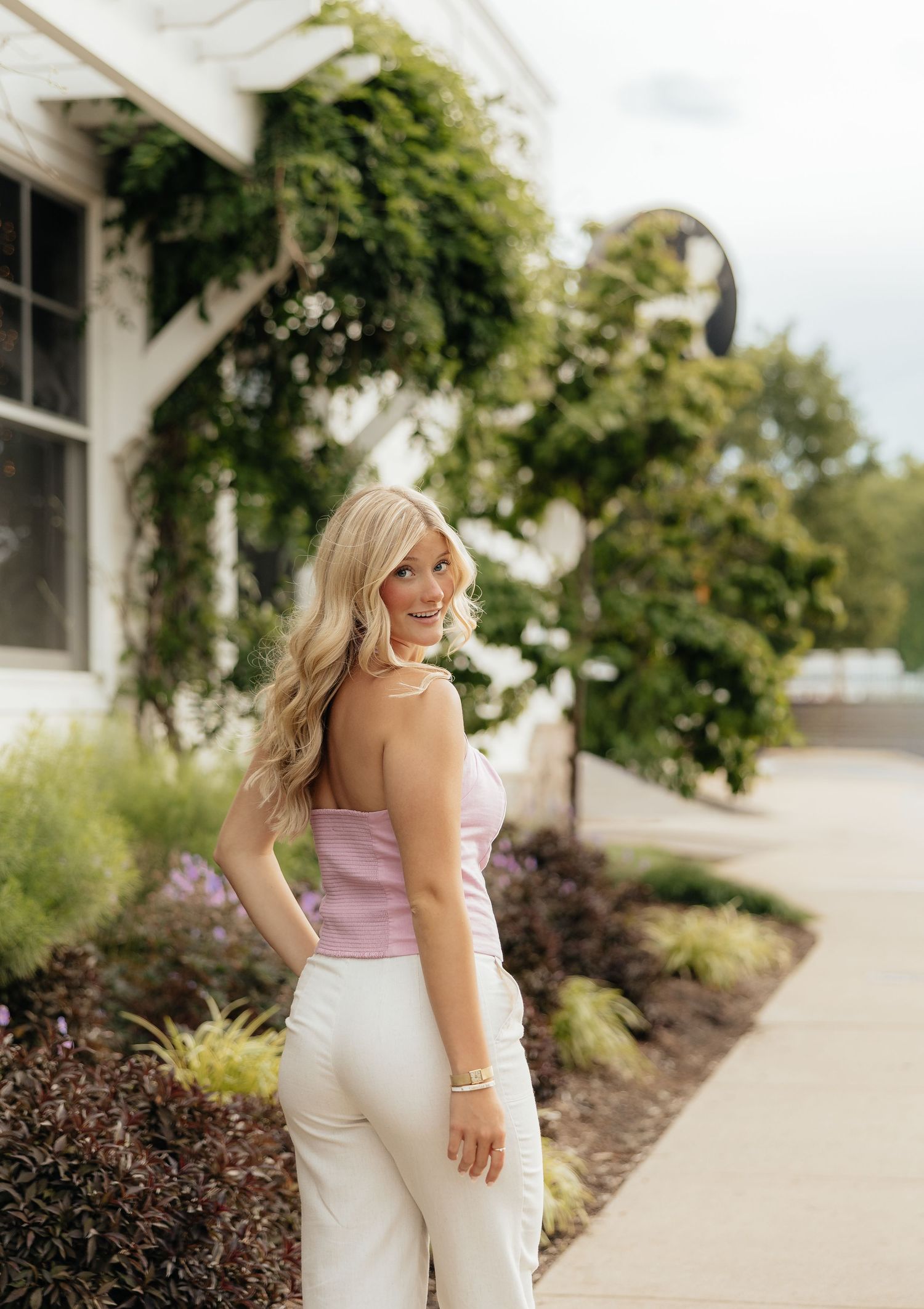 Blonde woman in pink top and white pants poses outdoors near ivy-covered building on a sunny day.