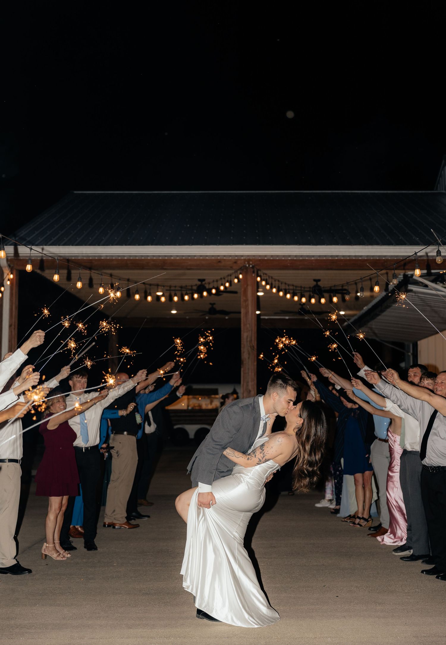Bride and groom share a romantic dip kiss during sparkler send-off exit at outdoor wedding reception at night.
