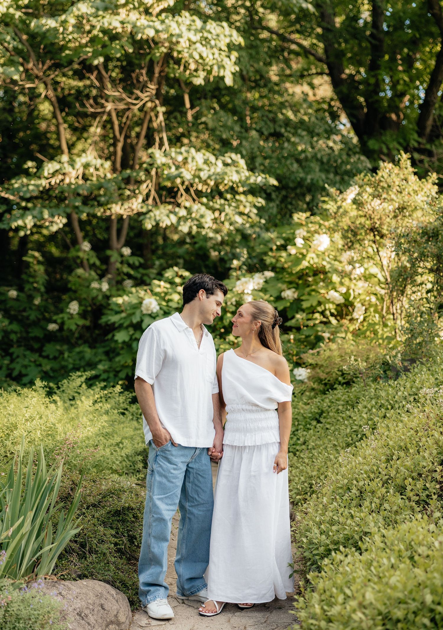 Couple in white outfits holding hands among lush green garden trees and shrubs on a sunny day.