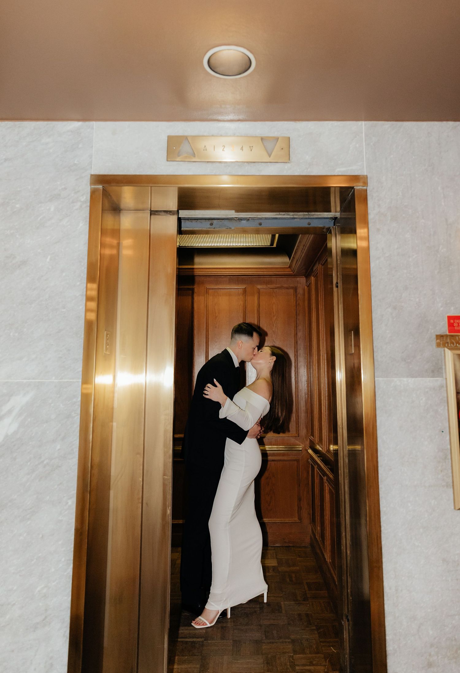 Elegant couple embraces in a golden-framed elevator doorway under soft lighting at an upscale venue.