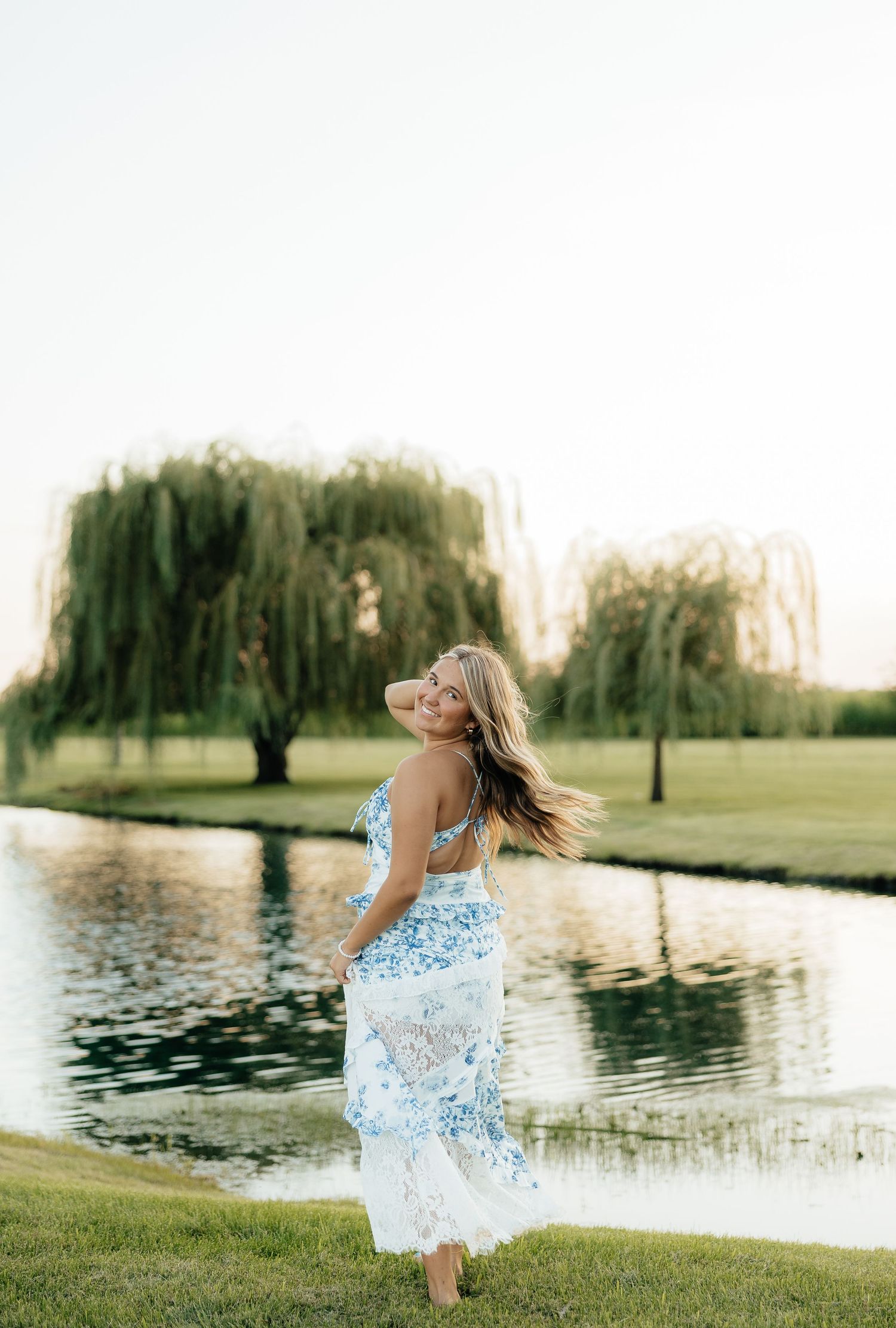 A woman in a blue floral dress twirls by a serene pond with weeping willow trees in golden hour light.