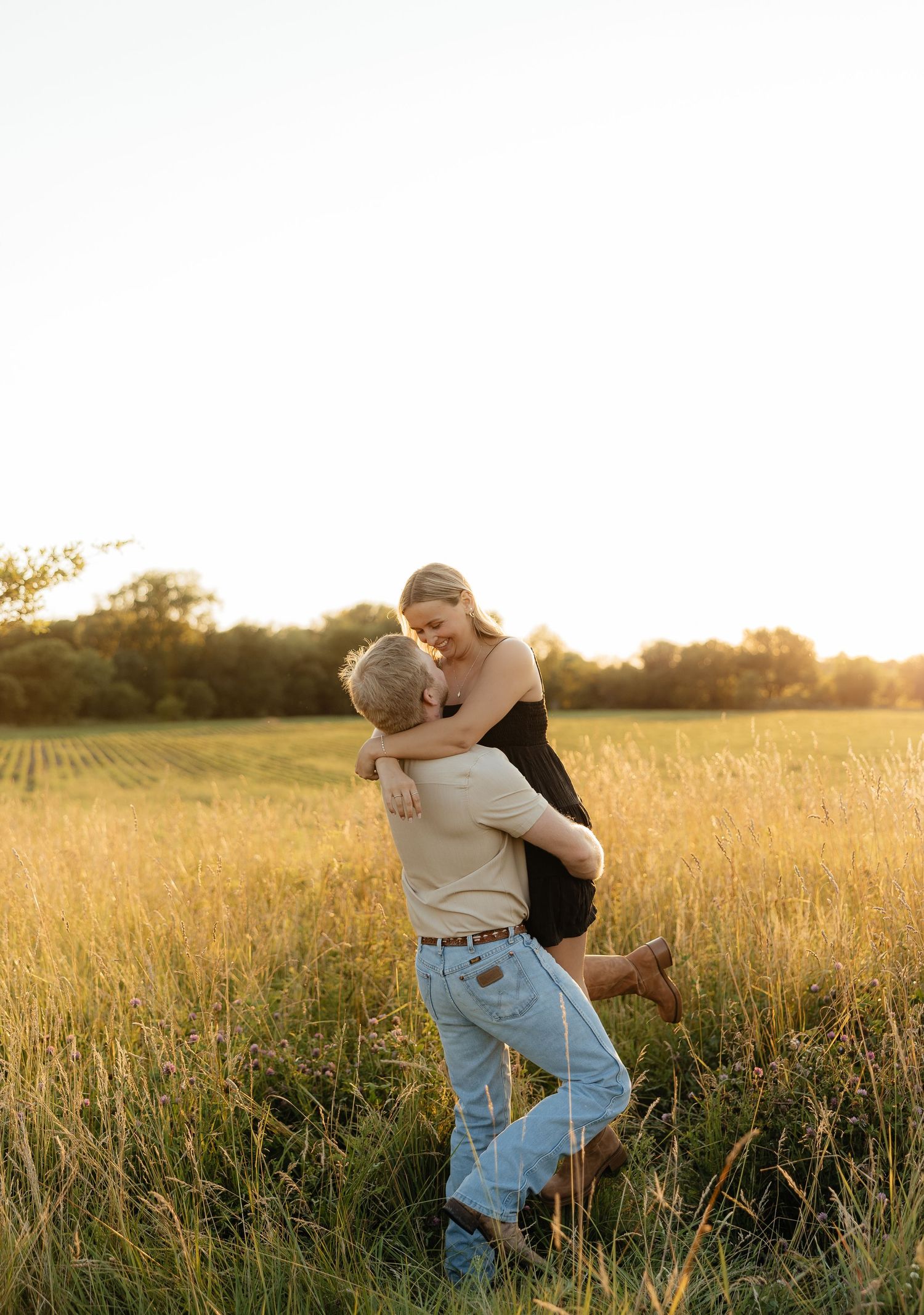 Couple embracing in golden hour sunlit field, man lifting woman in black dress among tall grass with trees in background.