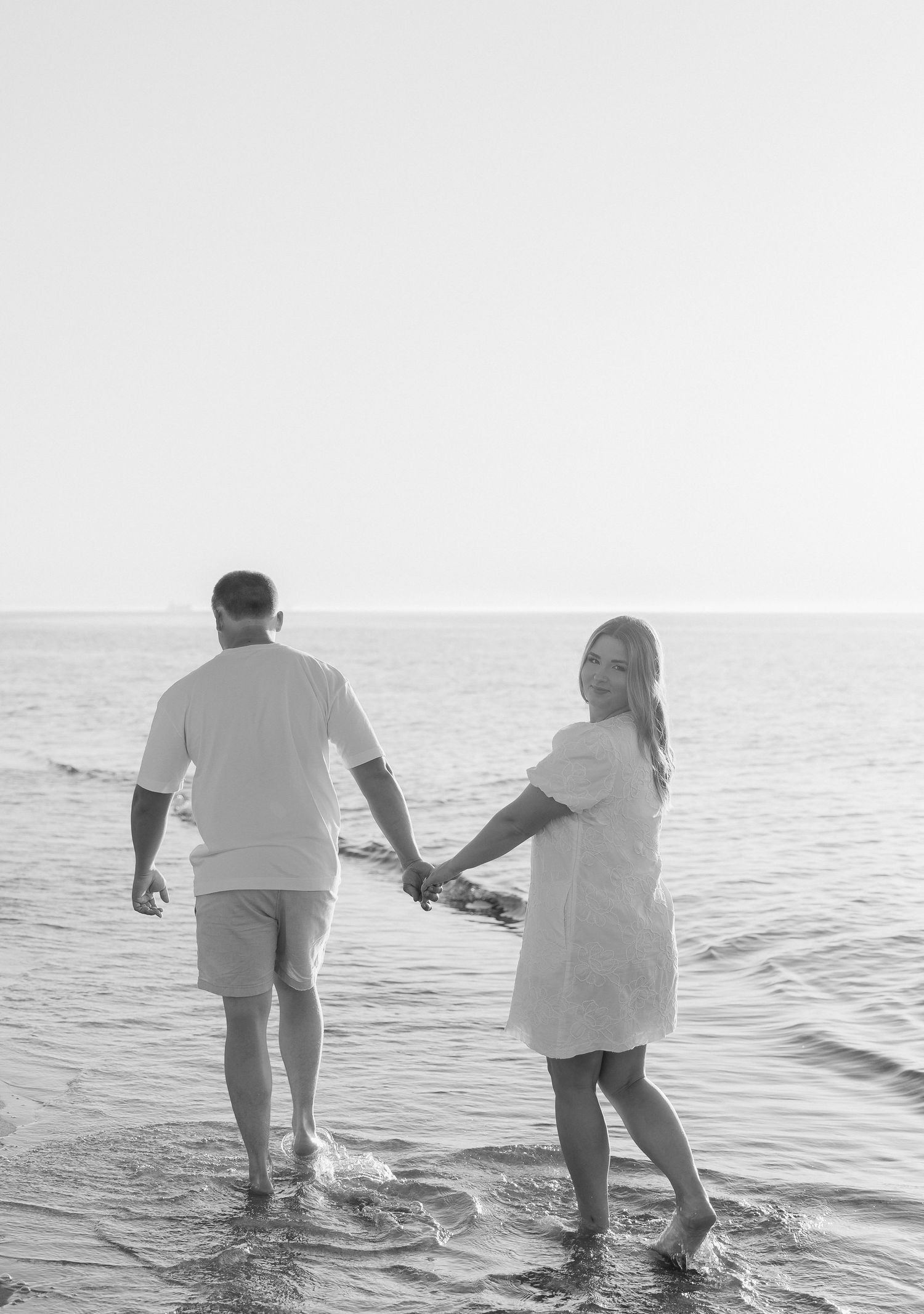 Couple holding hands walking in ocean waves on beach, black and white romantic photo.