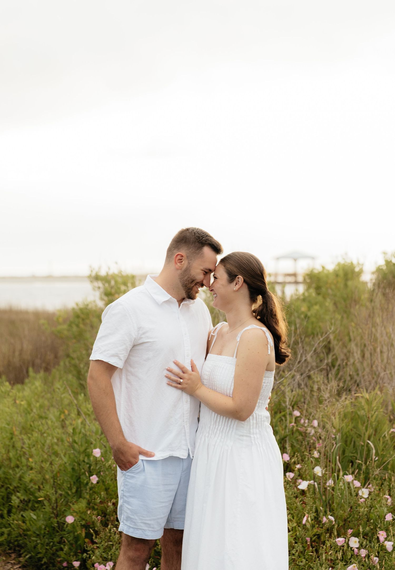 Couple in white outfits sharing a romantic moment surrounded by wildflowers and coastal greenery on a cloudy day.