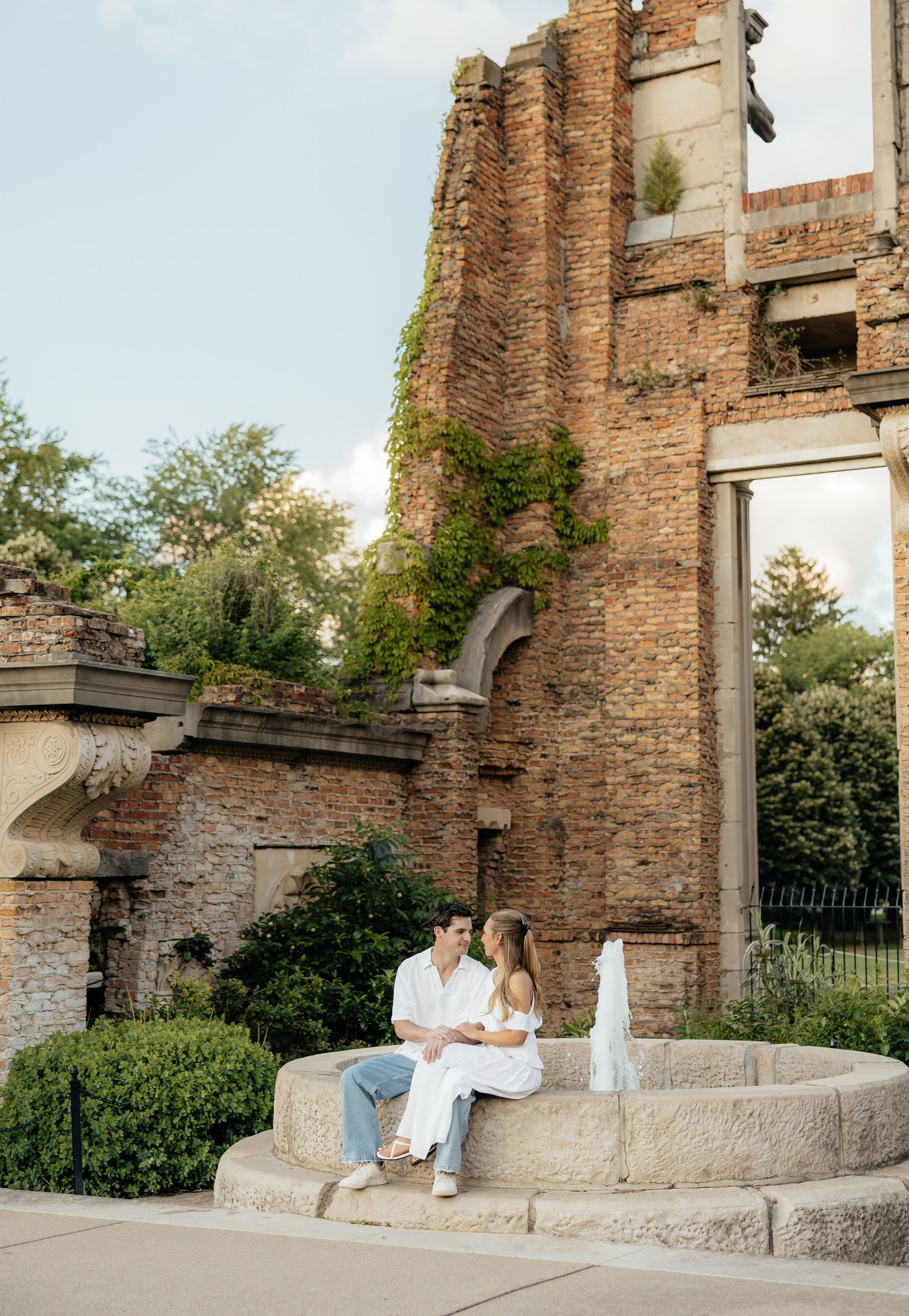 Couple sitting together on a stone fountain ledge in front of a rustic brick building with ivy, dressed in white outfits.