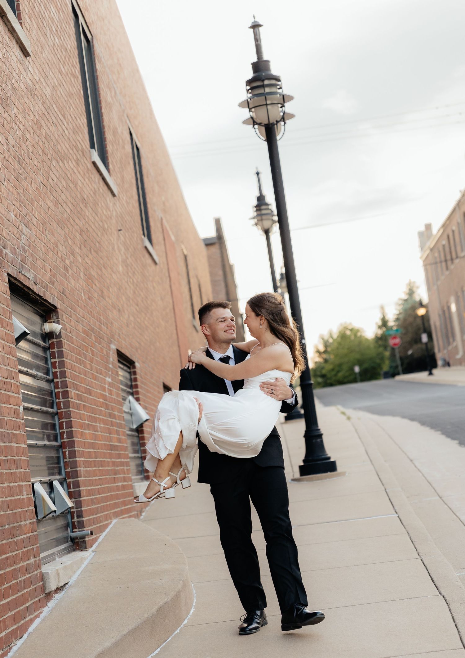 Groom carrying bride in his arms on a city sidewalk, both smiling joyfully in wedding attire.