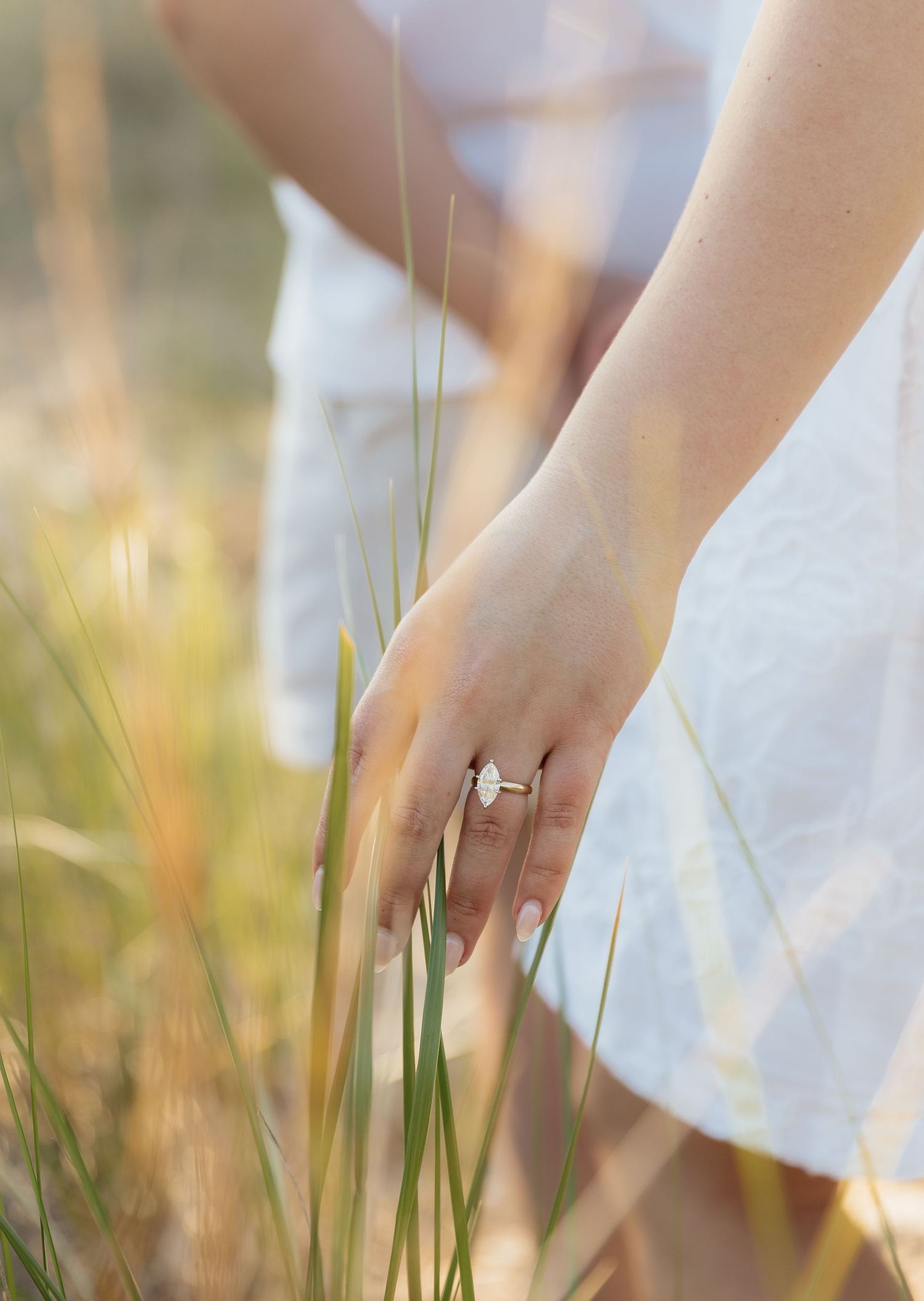 Close-up of a woman's hand wearing a diamond engagement ring, touching tall grass in a sunny field.