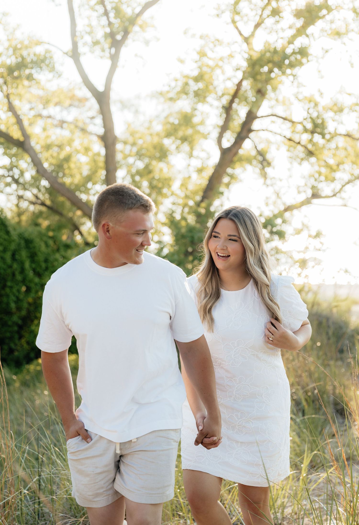 A smiling couple in matching white outfits holds hands while walking through a sunny, golden field with trees in the background.