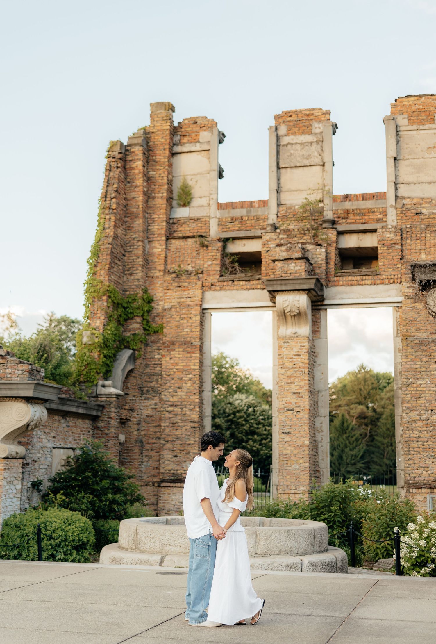 Couple in white dress and jeans embracing before rustic brick ruins with columns and lush greenery at golden hour.