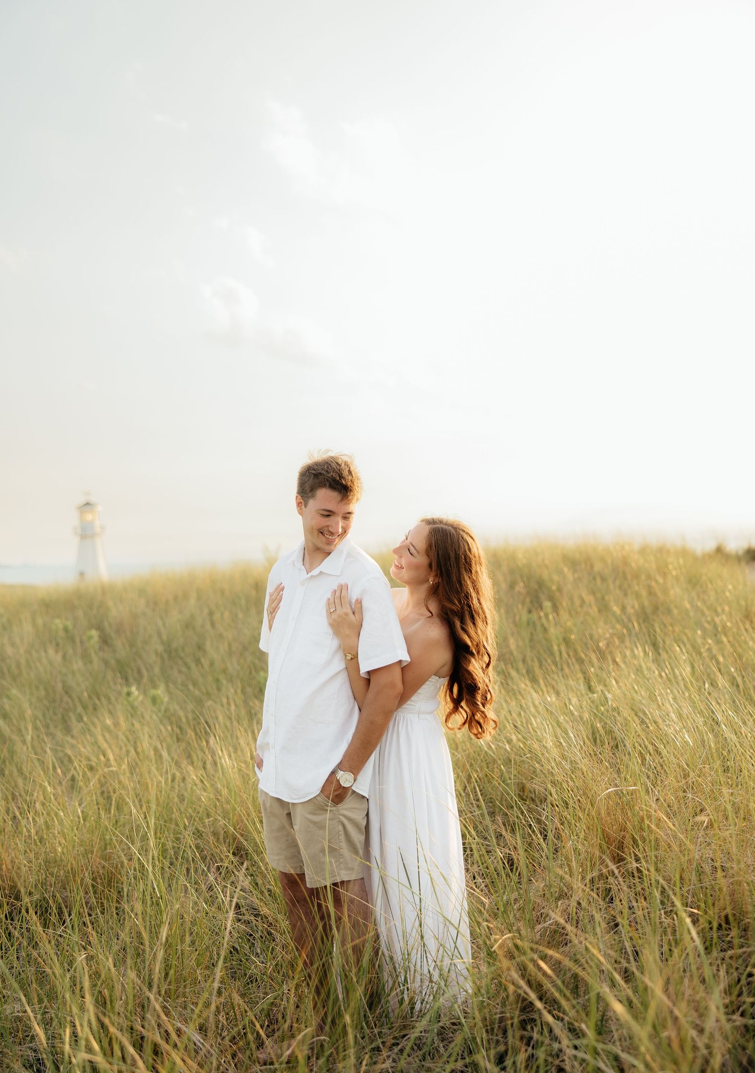 A couple in white outfits embracing in tall golden grass near a lighthouse on a bright sunny day.