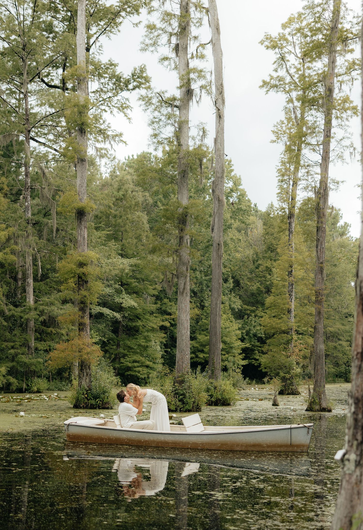 A couple shares a romantic kiss in a white boat on a serene, cypress-lined swamp, perfect for a wedding photo.