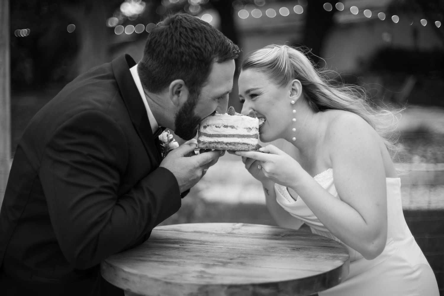 Black and white photo of bride and groom sharing a bite of wedding cake together at a small round table.