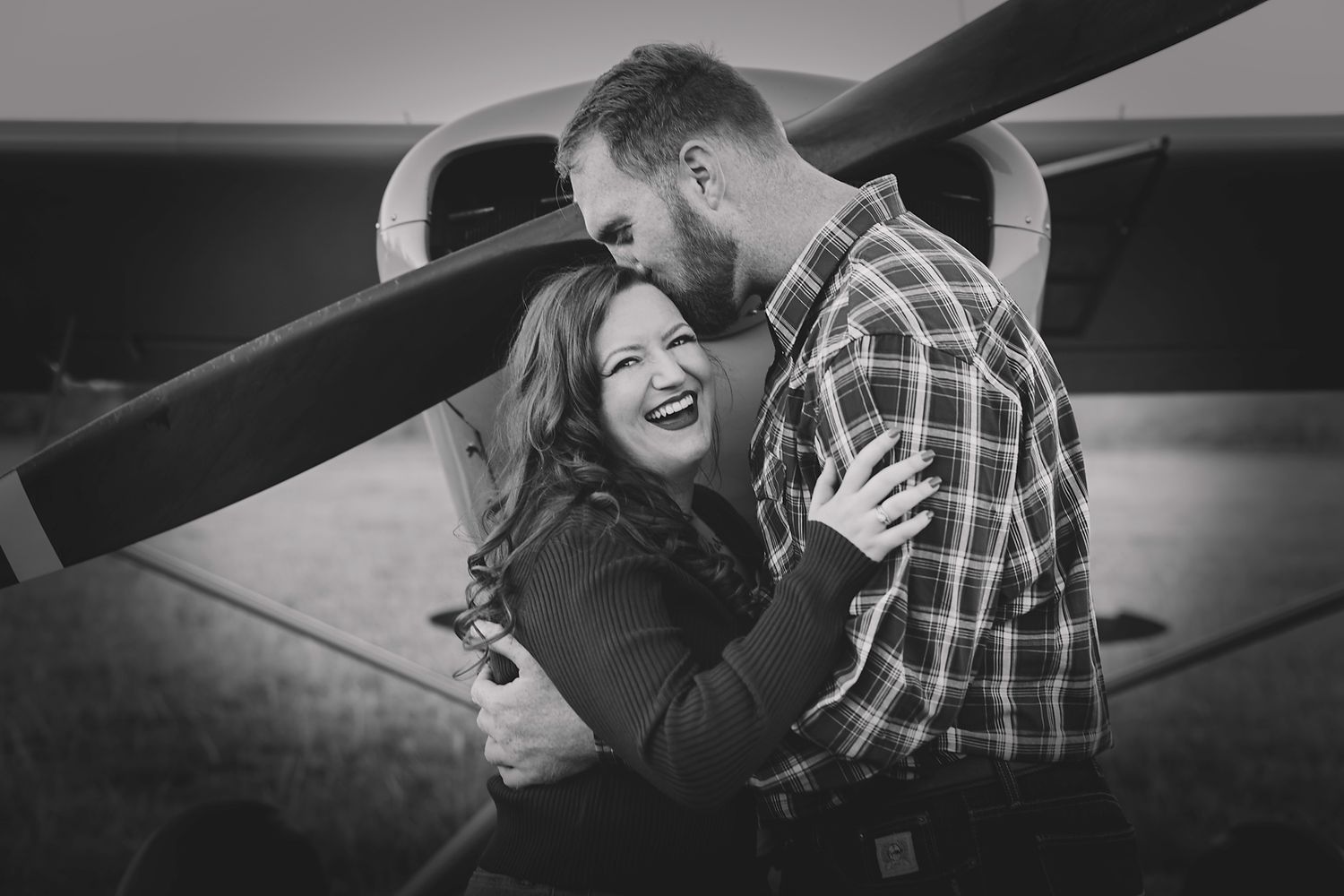 Black and white photo of a laughing couple embracing in front of a small propeller airplane outdoors.
