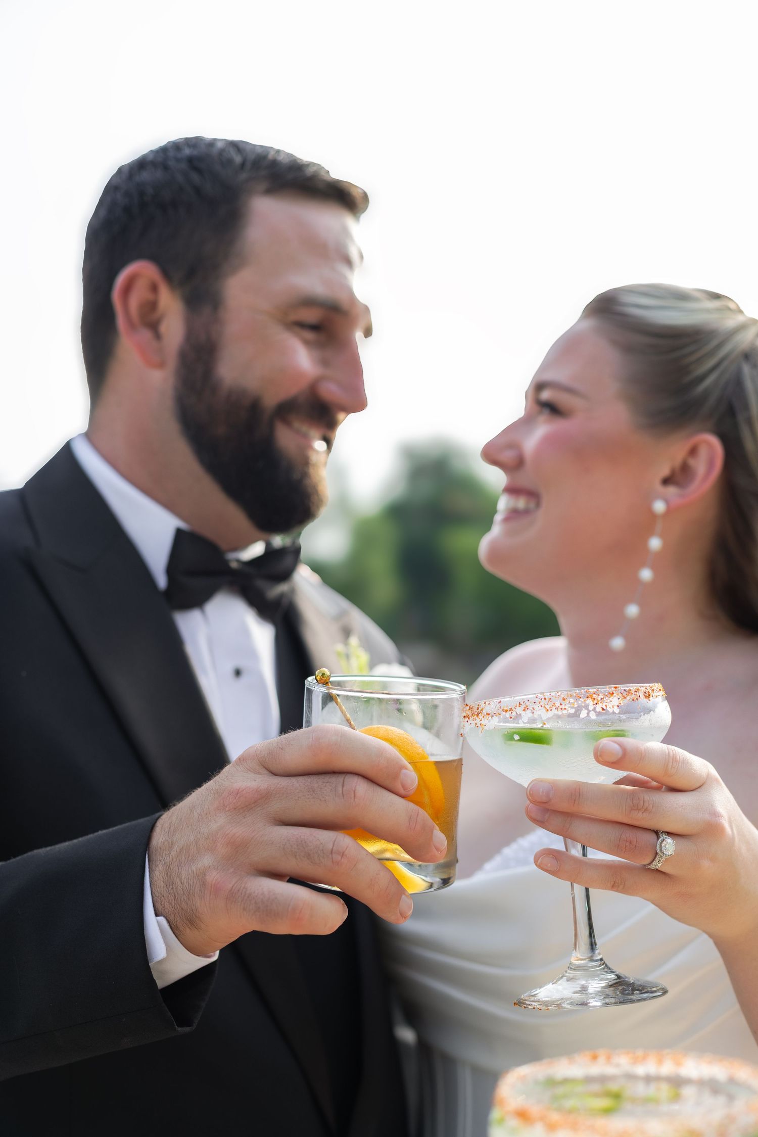Smiling bride and groom in formal attire clinking cocktail glasses together at their wedding reception outdoors.