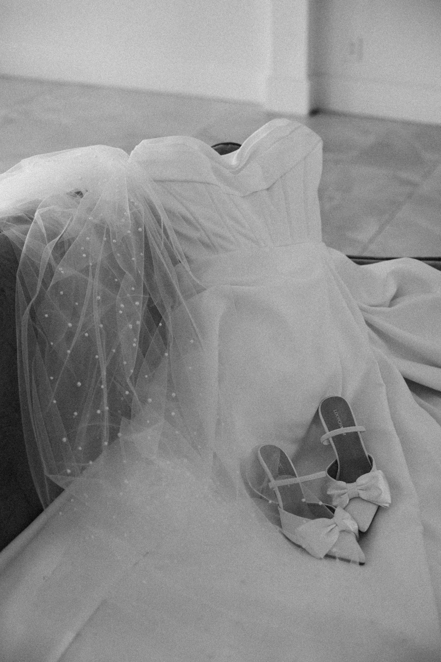 Black and white photo of a strapless wedding dress with dotted veil and bow heels on a sofa.