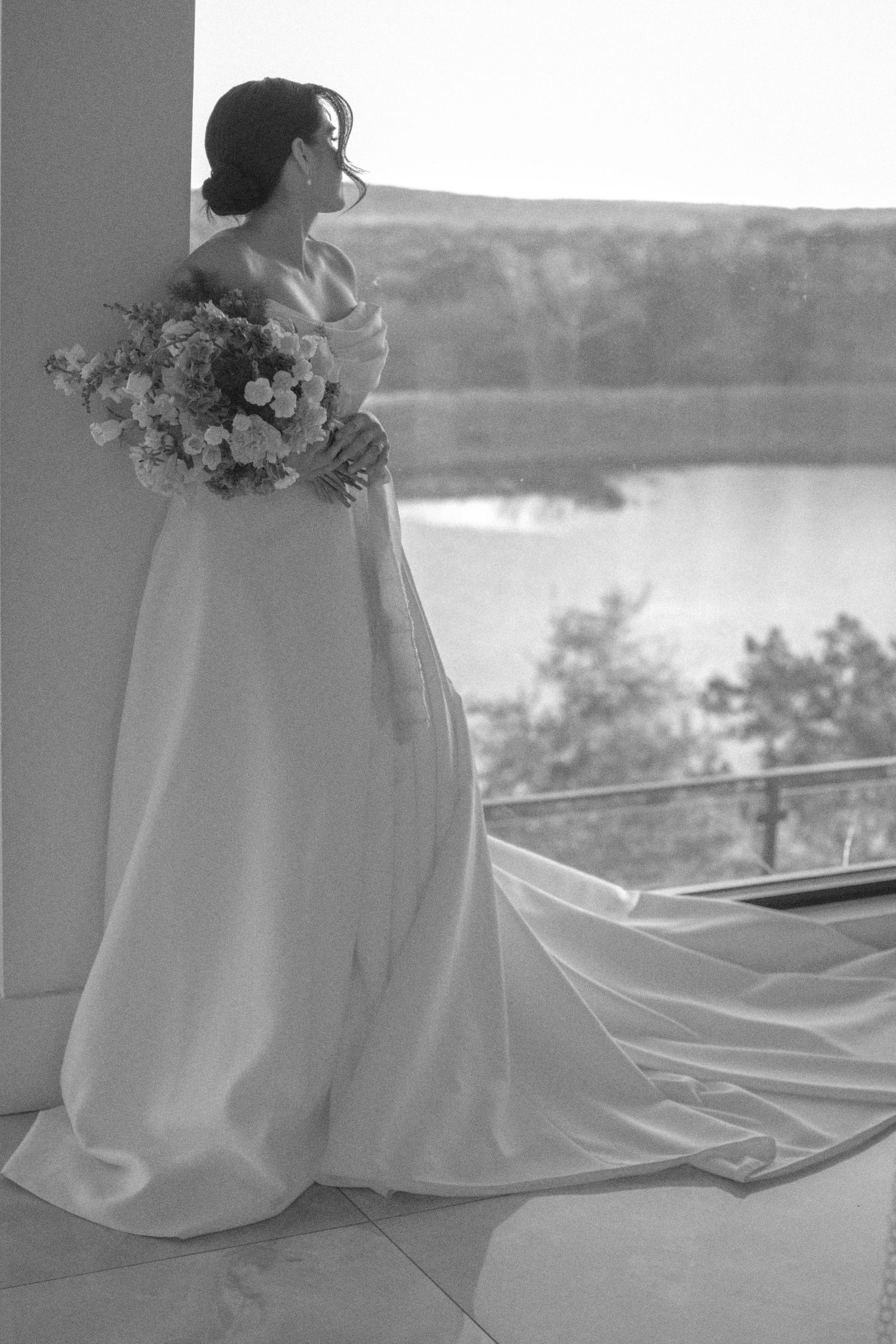 Black and white photo of a bride in an elegant gown with a long train, holding a bouquet by a window overlooking a lake.