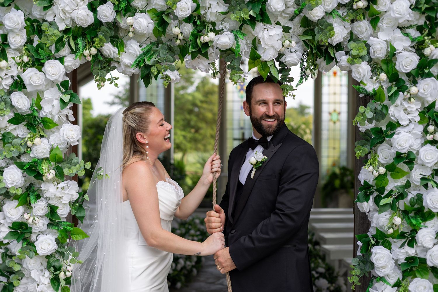 Bride and groom laughing together under a white floral arch adorned with greenery at their wedding ceremony.