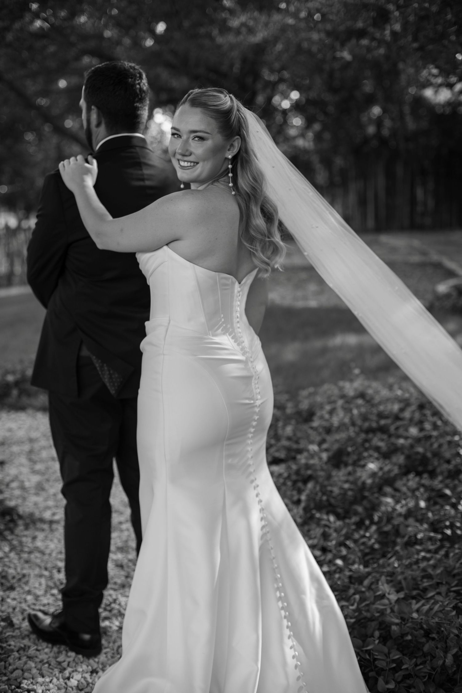 Bride in fitted white gown and veil smiles over shoulder at groom in black tuxedo outdoors in black and white photo.