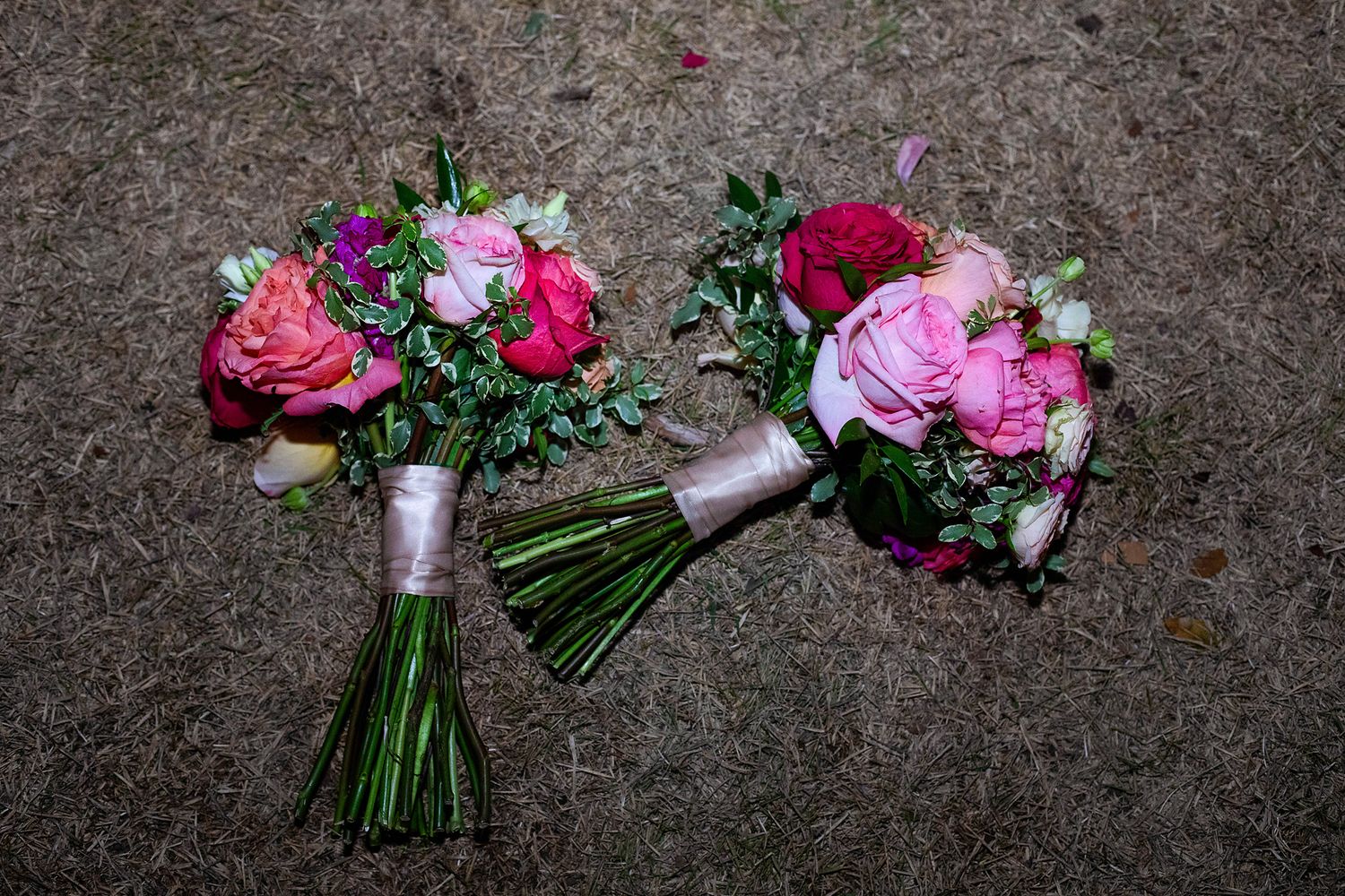 Two vibrant pink and red wedding bouquets with greenery, tied with ribbon, resting on a textured gray surface.