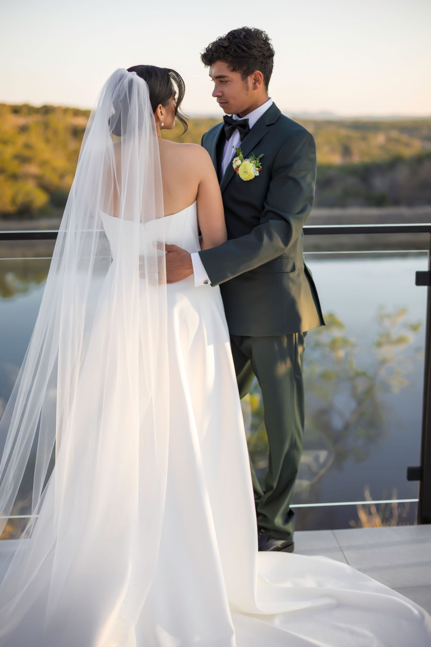 Bride in white gown and veil embraces groom in dark tuxedo on outdoor terrace with scenic water view.