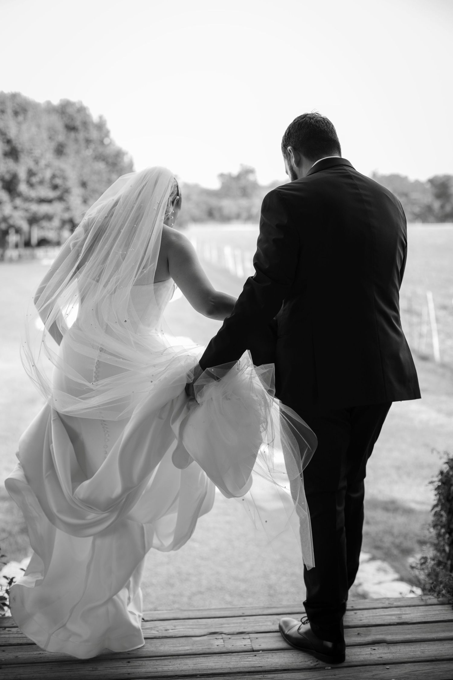 Black and white photo of bride and groom walking hand in hand, her veil flowing in the breeze on their wedding day.