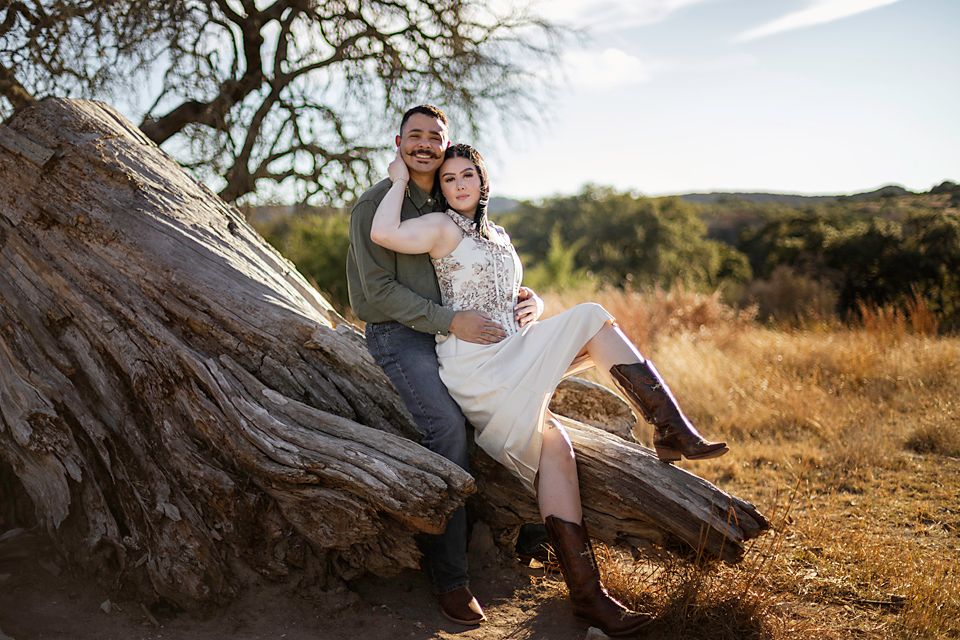 Couple posing on a rustic log in a golden field, woman in white dress and boots, man in green shirt, outdoor engagement photo.