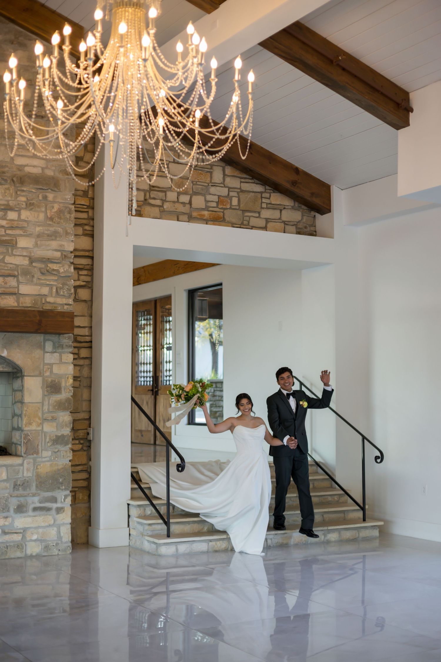Bride and groom descend elegant staircase in wedding venue with chandelier and stone fireplace.