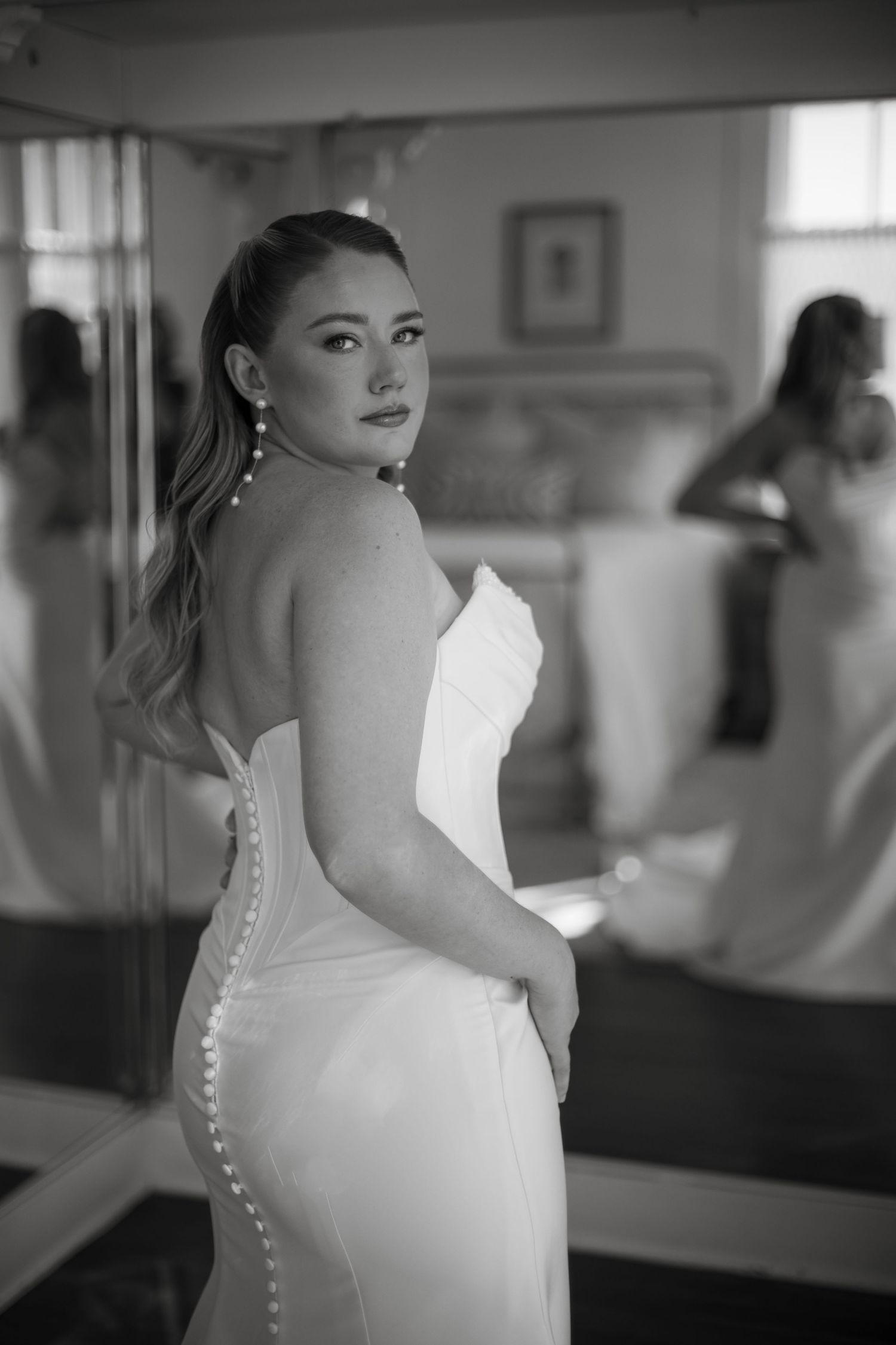 Black and white photo of a bride in a strapless gown with pearl buttons, posing elegantly in front of a mirror.