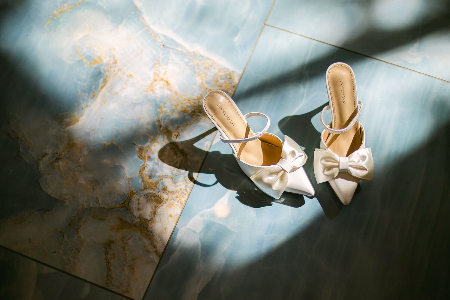 Elegant white satin bridal heels with bow detail casting shadows on marble floor in natural light.