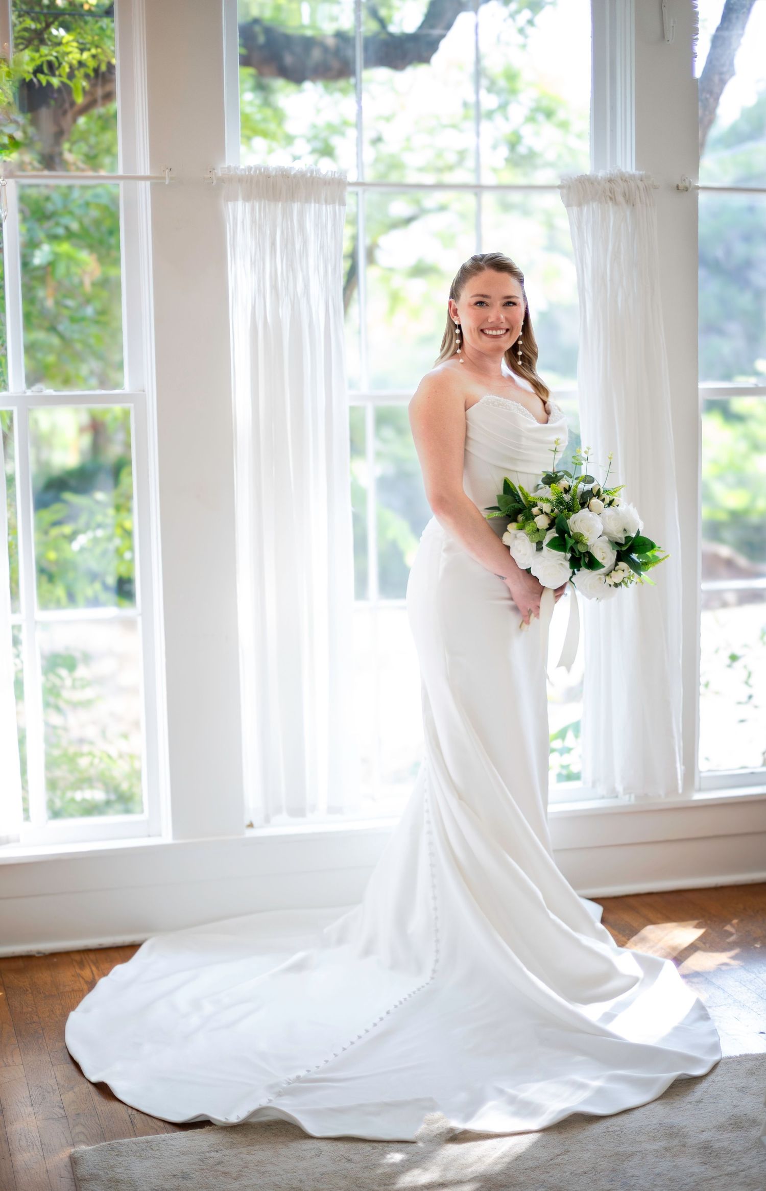 Bride in white satin gown with train holds bouquet of white roses, standing before large windows with sheer curtains.