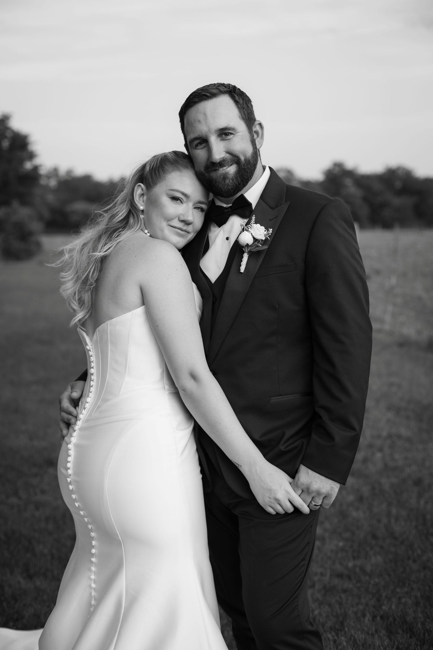 Black and white photo of a smiling bride in strapless gown and groom in tuxedo posing outdoors on their wedding day.