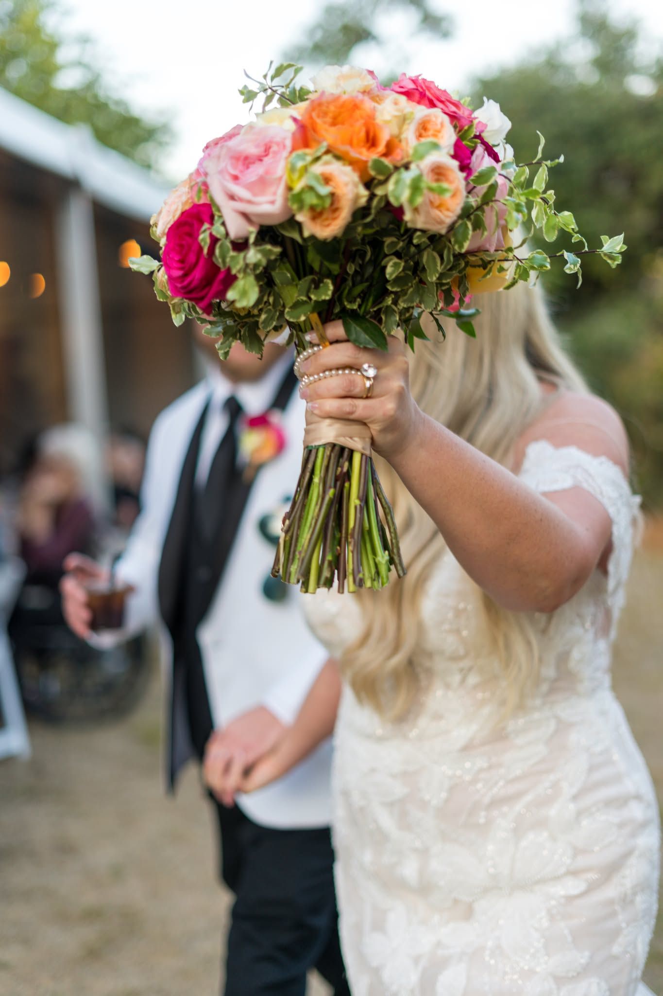 Bride in lace gown holds vibrant pink, orange, and coral rose bouquet with groom in black tie behind her.