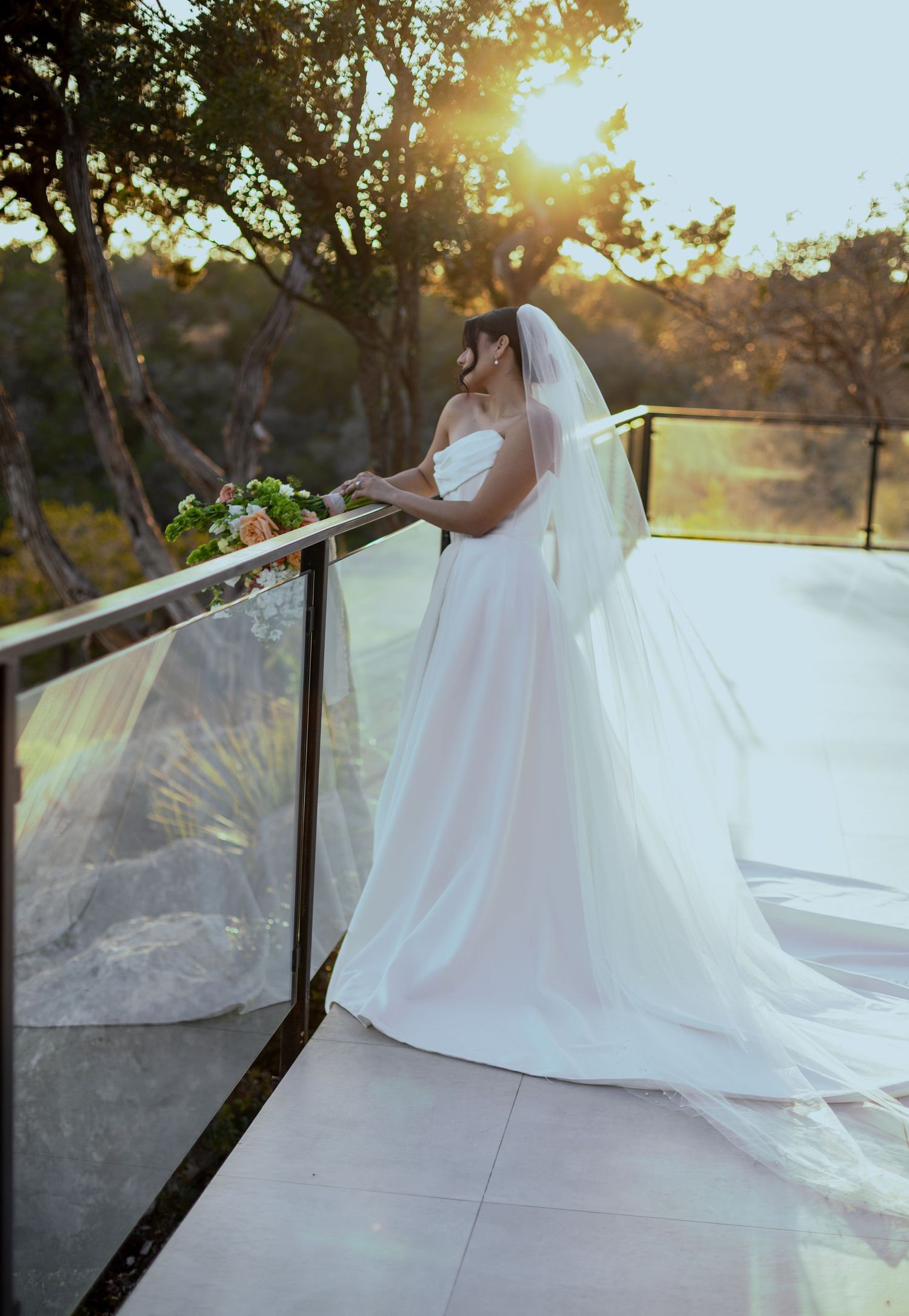 Bride in a white strapless gown and veil holds bouquet on sunlit balcony surrounded by trees at golden hour.