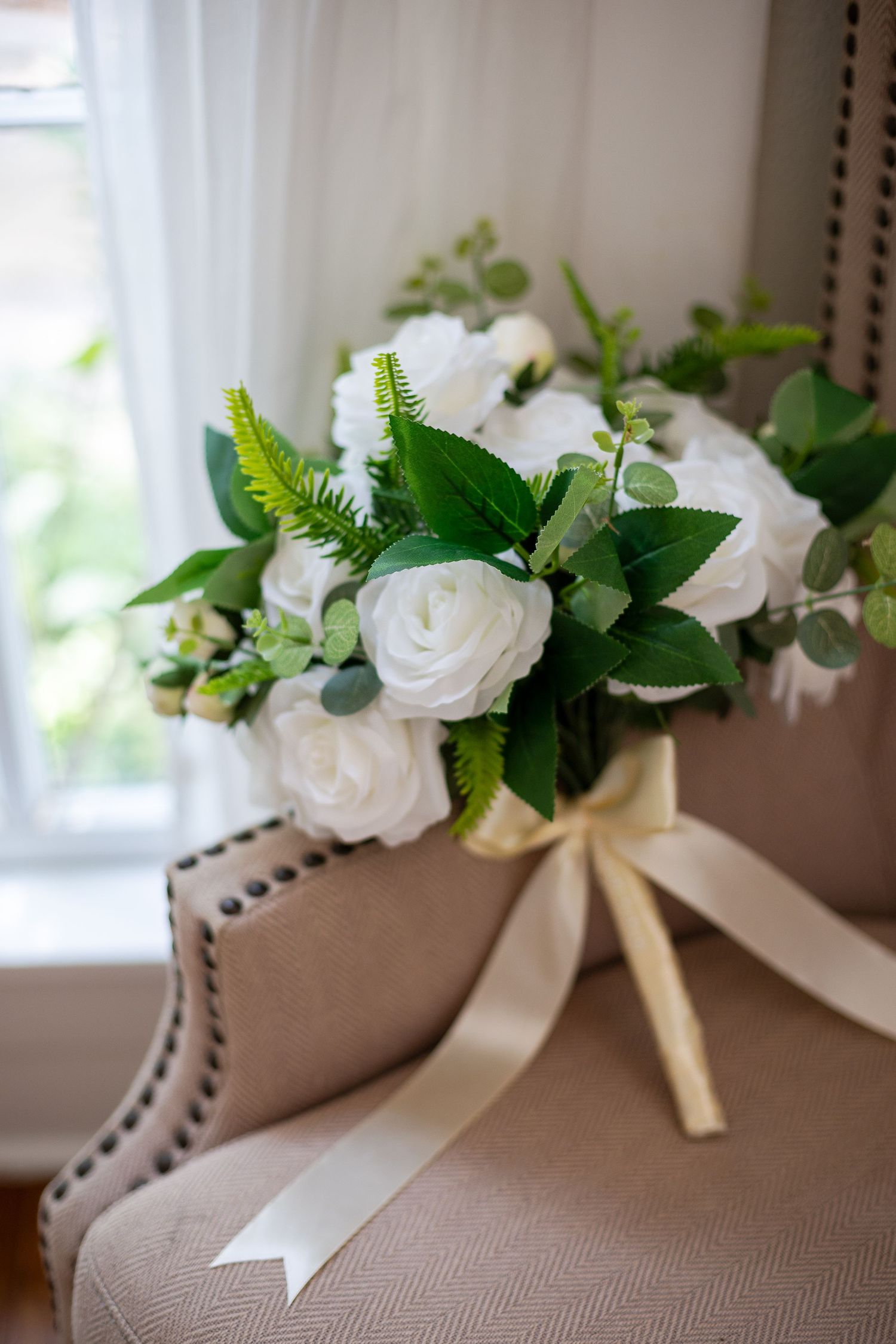 Elegant bridal bouquet of white roses and green foliage tied with cream ribbon, resting on a beige armchair.