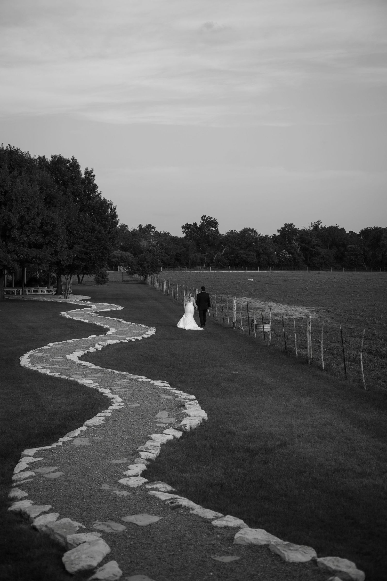 Black and white photo of a bride and groom walking along a winding stone path through a lush green field.