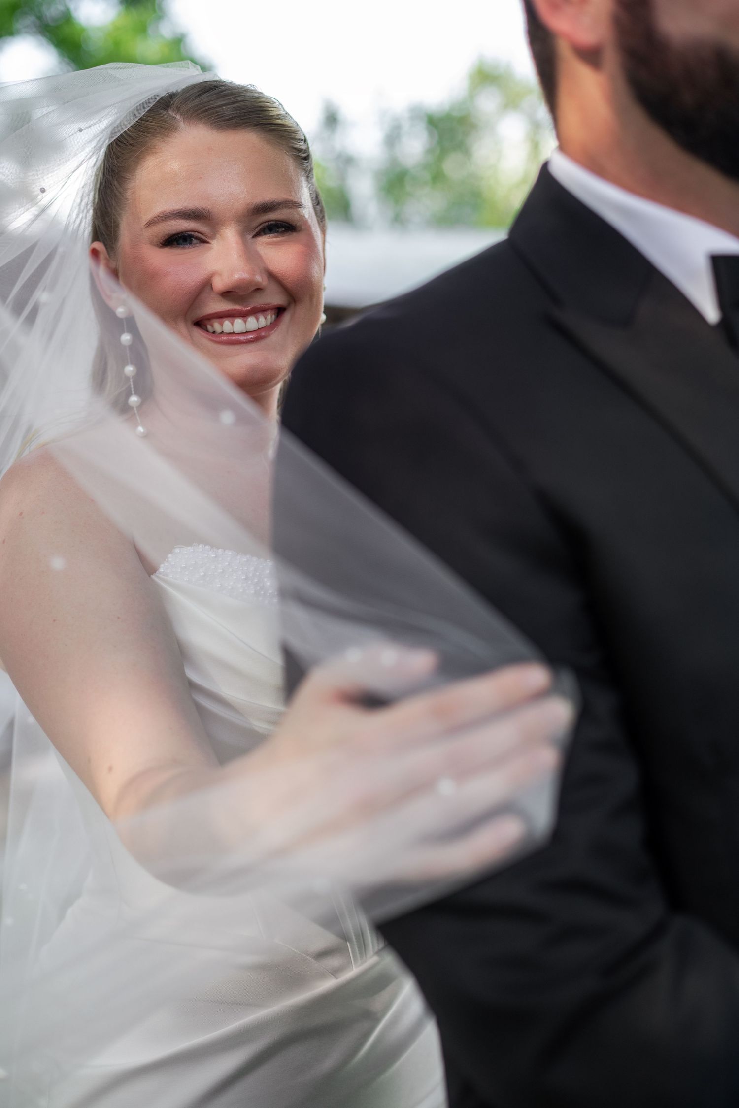 Smiling bride in white gown and veil holds groom in black suit during outdoor wedding ceremony.