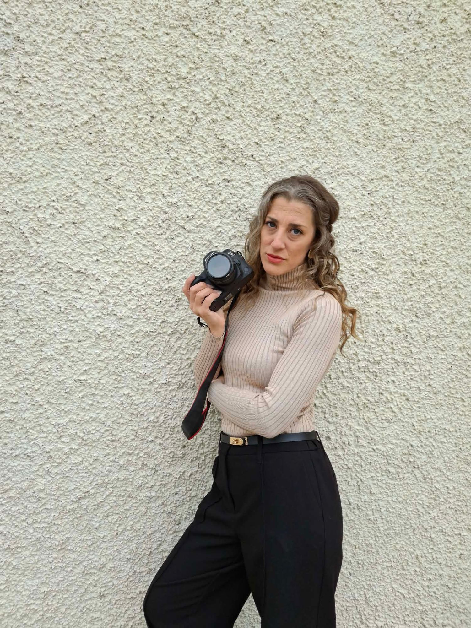 Woman in beige turtleneck and black trousers holding a camera against a white textured wall.