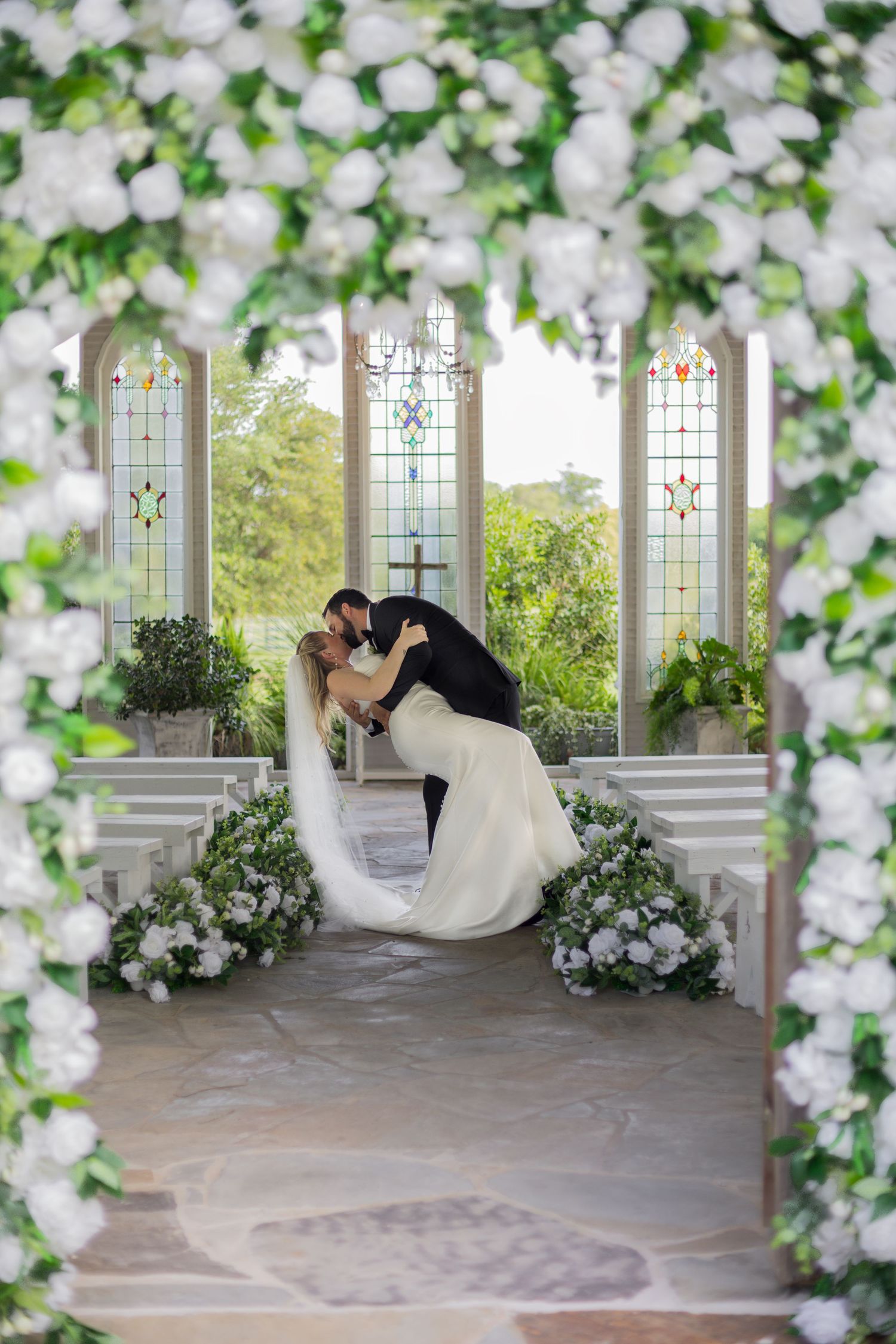 Bride and groom share a romantic dip kiss at the altar of a white chapel adorned with lush white floral arrangements.