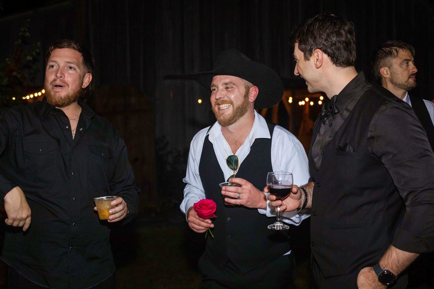 Three men in black attire laugh and socialize at an evening event, one holding a red rose and wine glass.