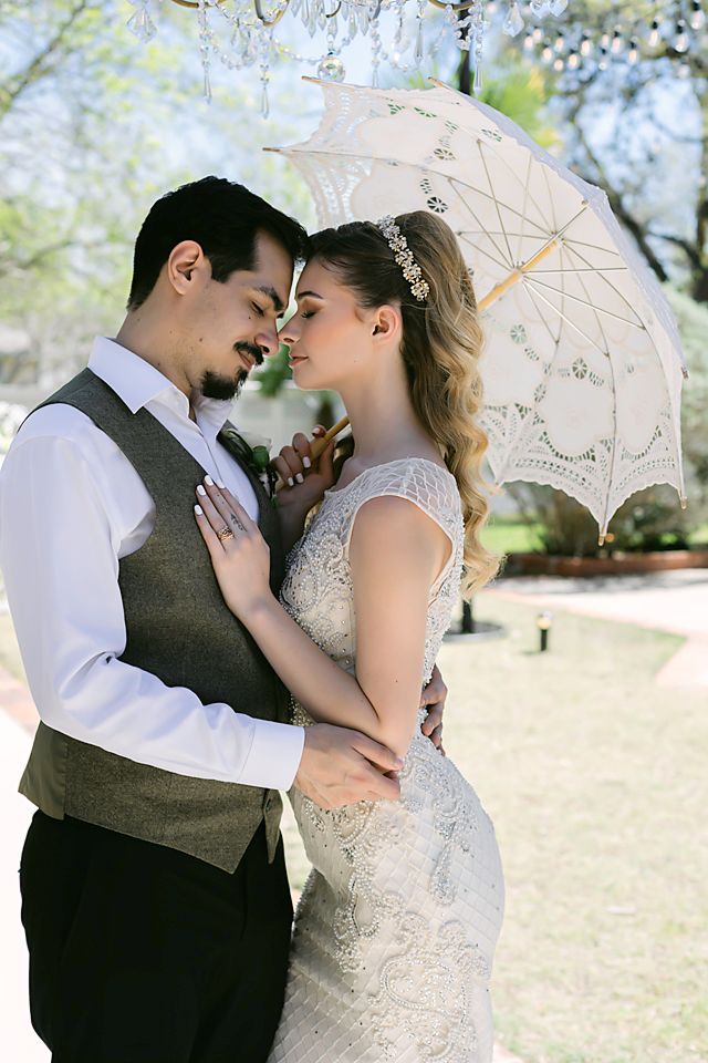 Bride and groom share a romantic moment under a lace parasol outdoors in a sunlit garden on their wedding day.