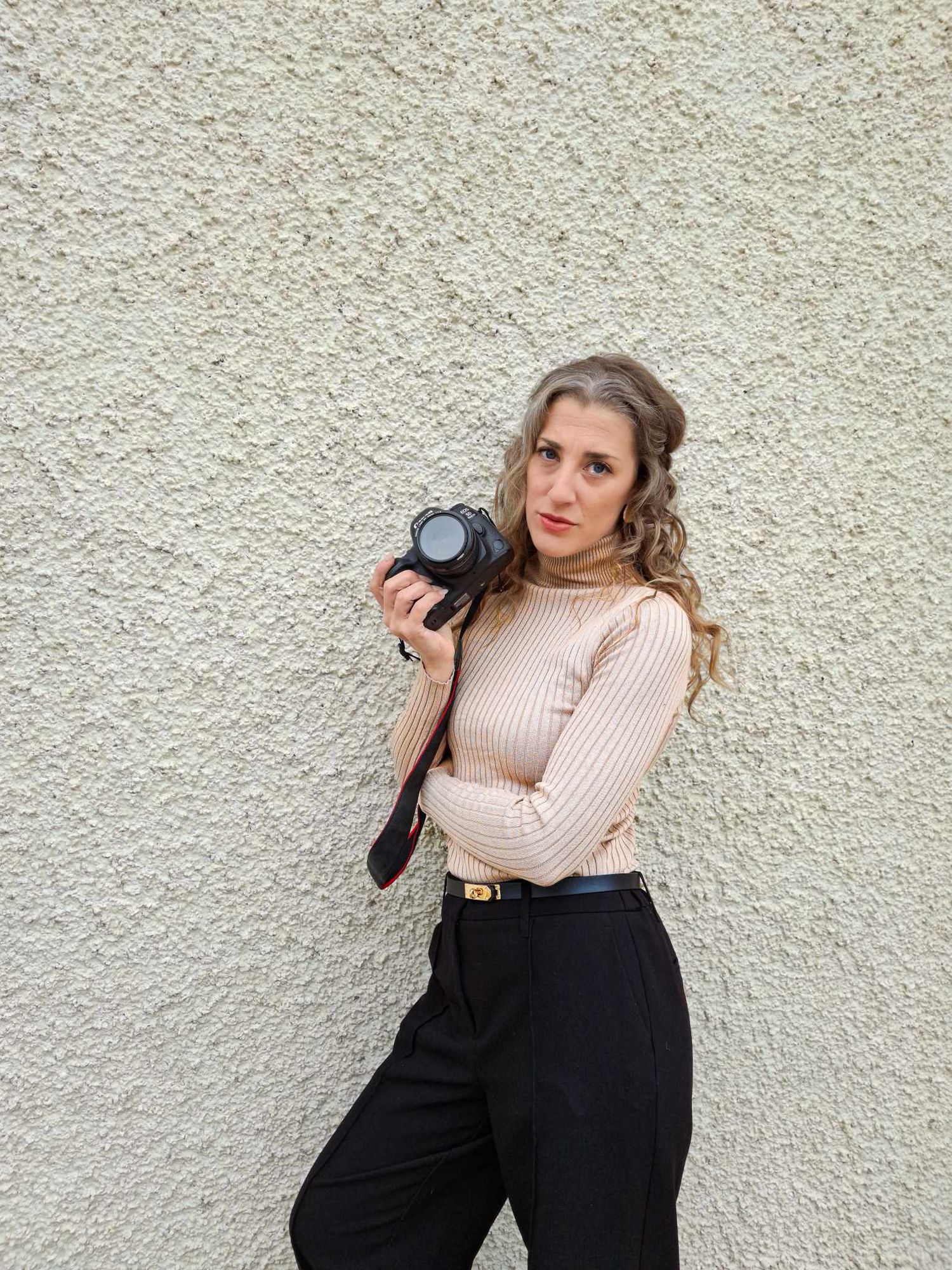 Young woman in beige turtleneck and black trousers holding a DSLR camera against a textured white wall.
