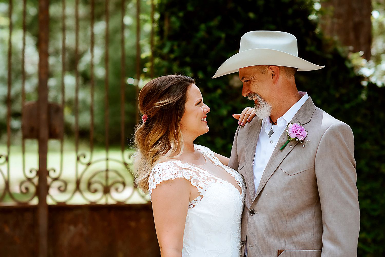 Smiling bride and groom in a tan suit and cowboy hat share a joyful moment outdoors at their wedding.