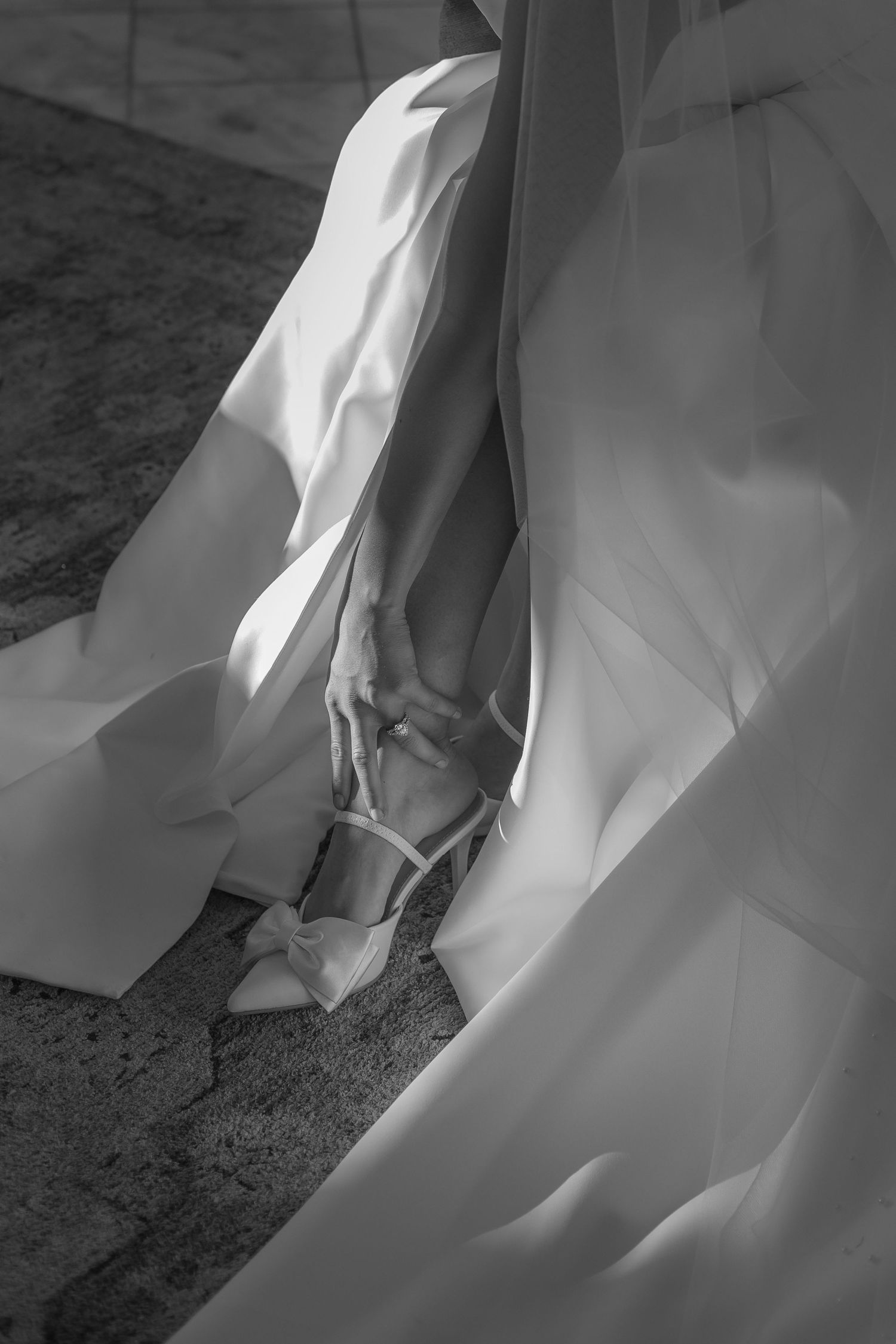 Black and white photo of a bride adjusting her elegant bow heels beneath a flowing white wedding gown.