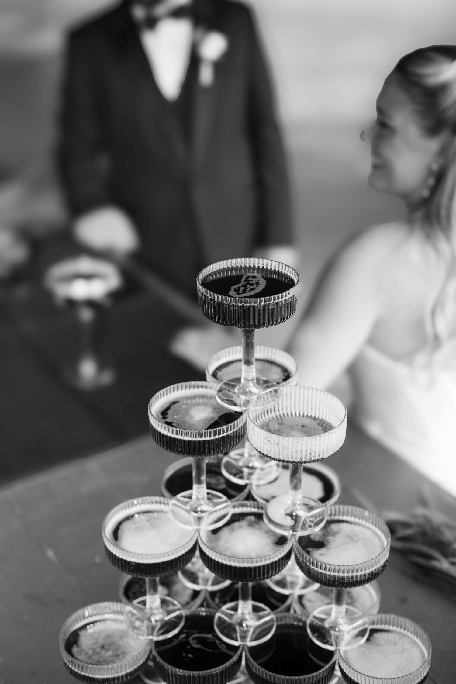 Black and white photo of a champagne tower at a wedding reception with bride and groom in the background.